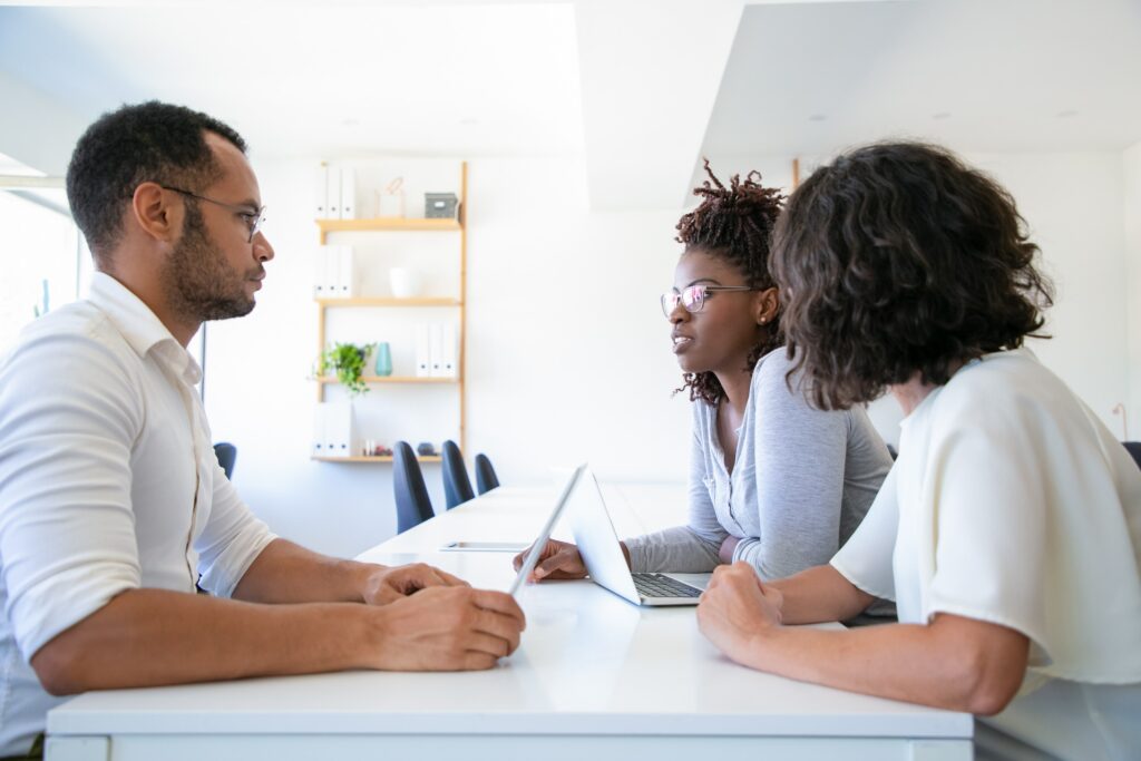 Duas mulheres e um homem sentados frente a frente em uma mesa de reunião, onde há dois laptops.