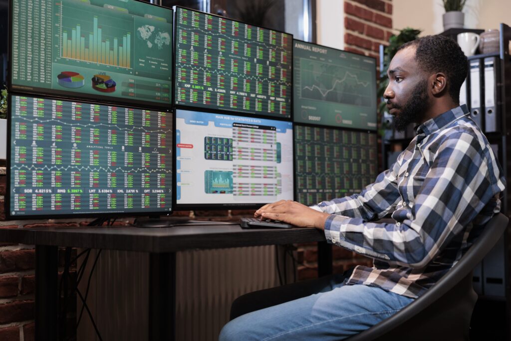 Homem negro sentado em frente a várias telas observando ativos financeiros.