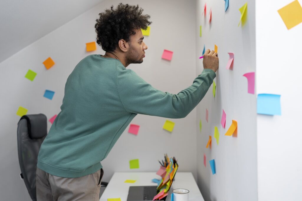 Homem escrevendo em um post-it laranja em uma parede cheia de outros post-its.