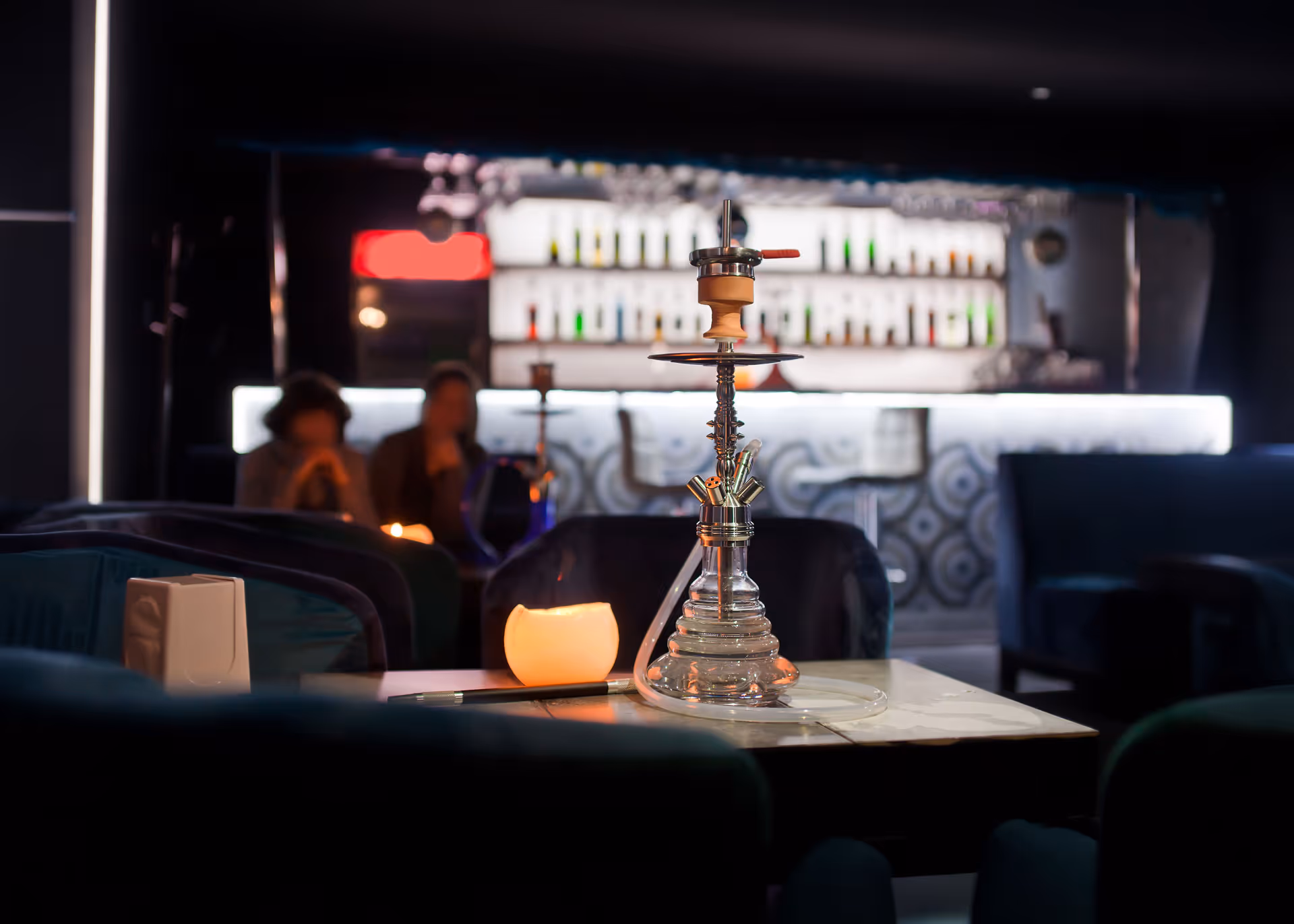 A hookah pipe on a table with a lit candle in a dim lounge with blurred people and bar shelves in the background.