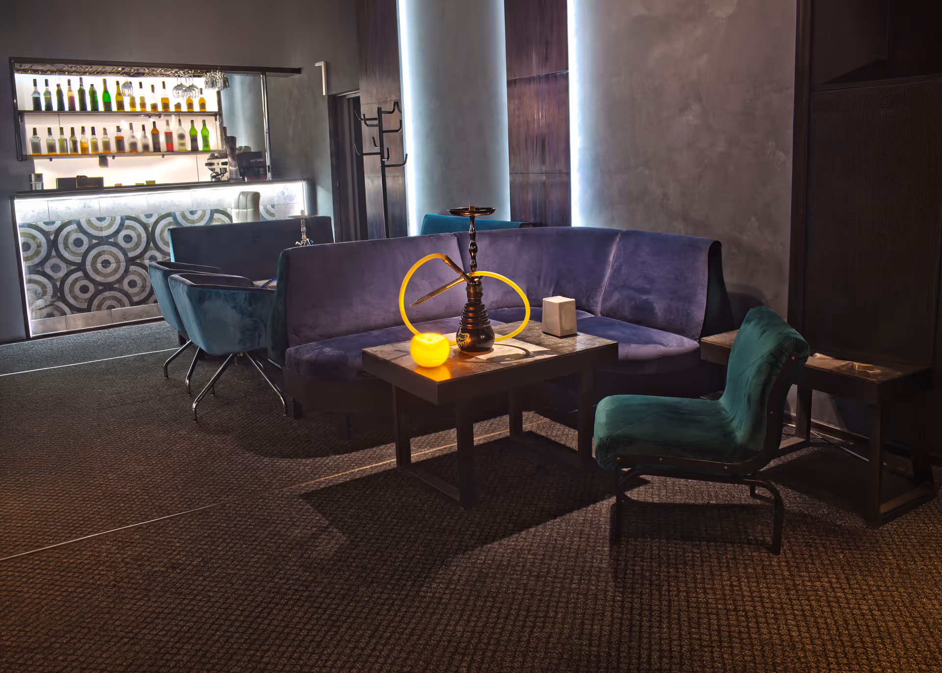 Dim lounge with purple velvet sofas, green chairs, a hookah on a wooden table, and backlit bar bottles.
