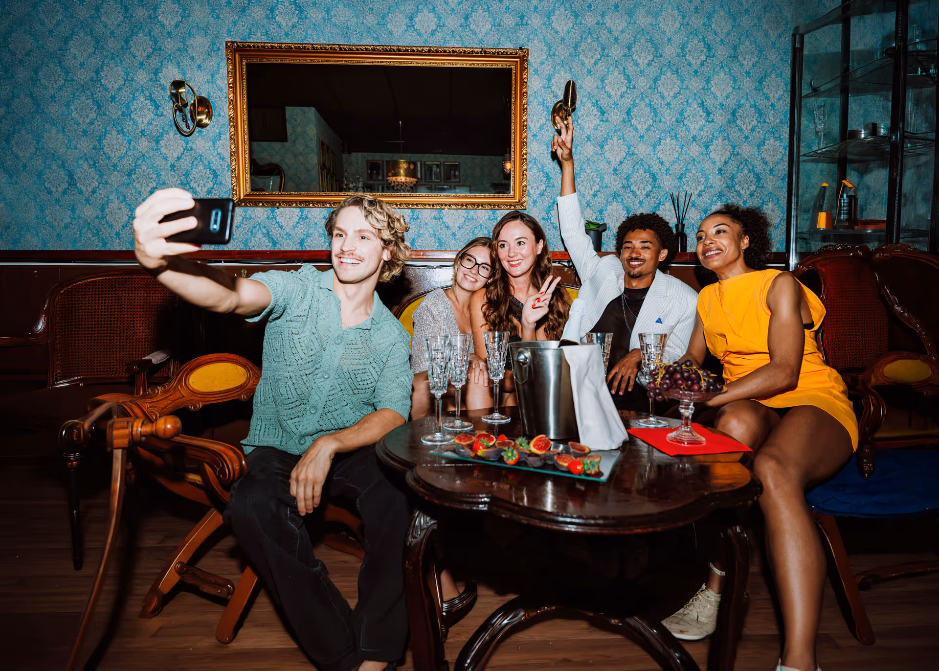 Five friends sitting in vintage chairs, taking a selfie with drinks on the table.