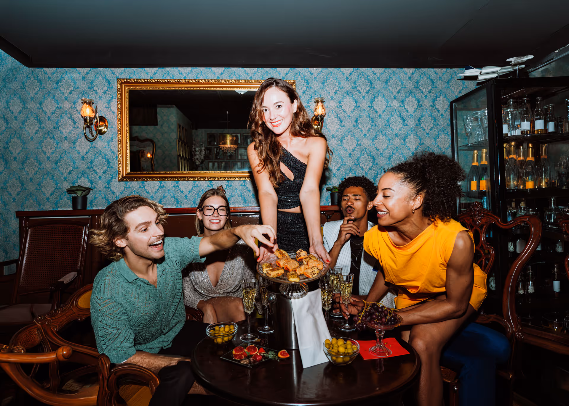 Five friends enjoy drinks and snacks in a vintage room; a woman offers a plate of appetizers.