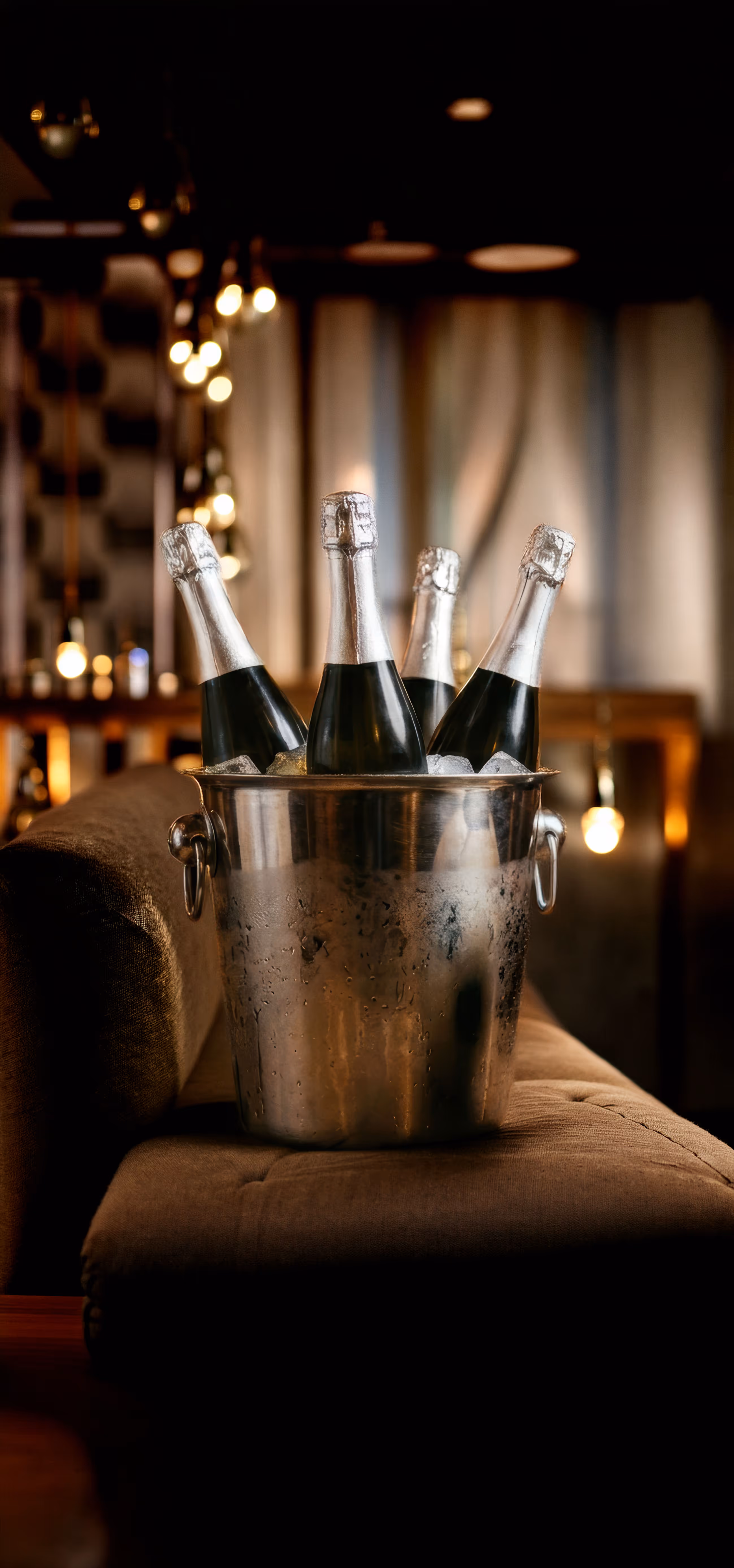 Four bottles with foil tops chilling in an ice-filled metal bucket on a brown cushioned surface.