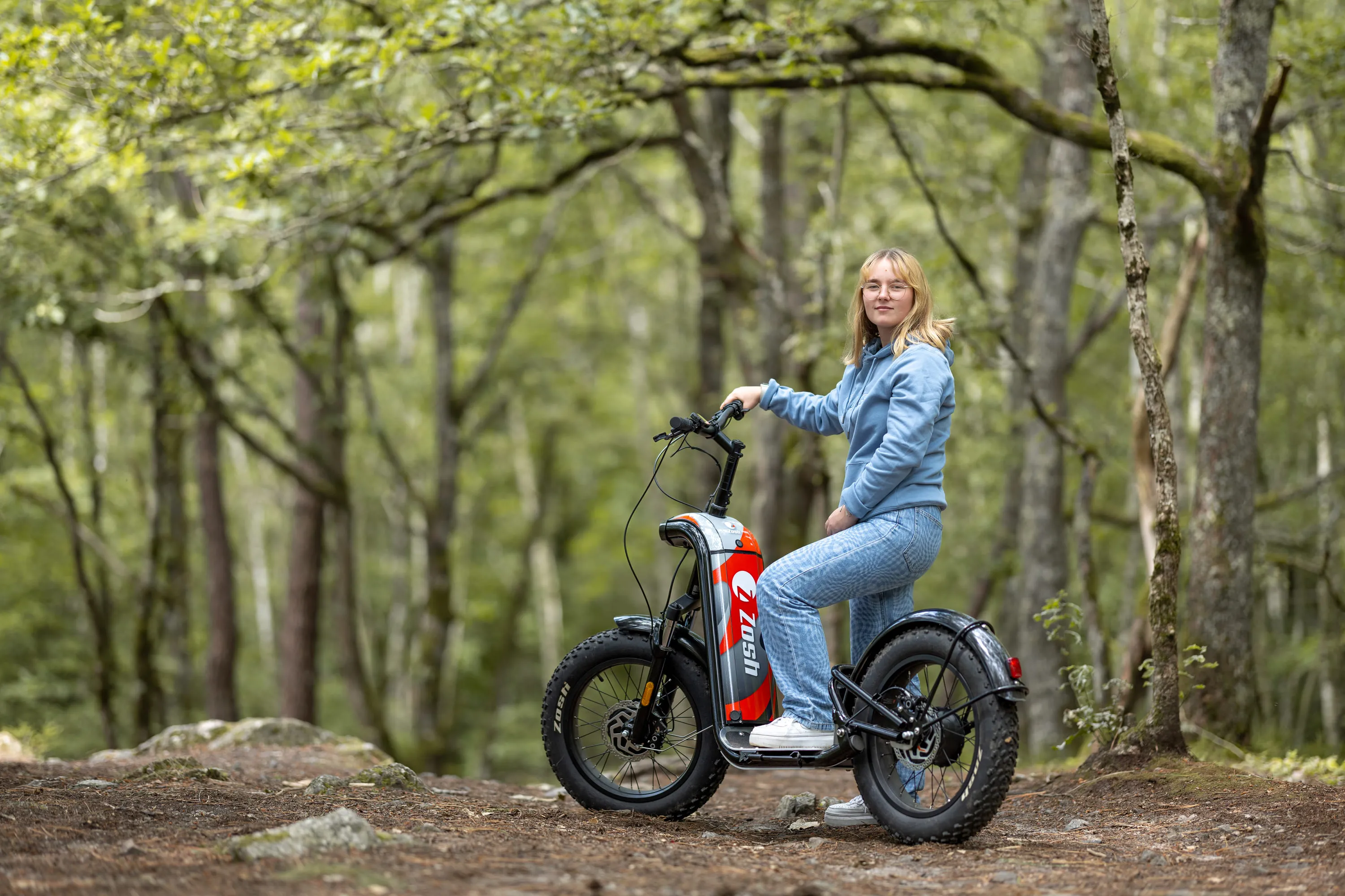 Jeune femme en sweat bleu sur une trottinette électrique tout-terrain en forêt.