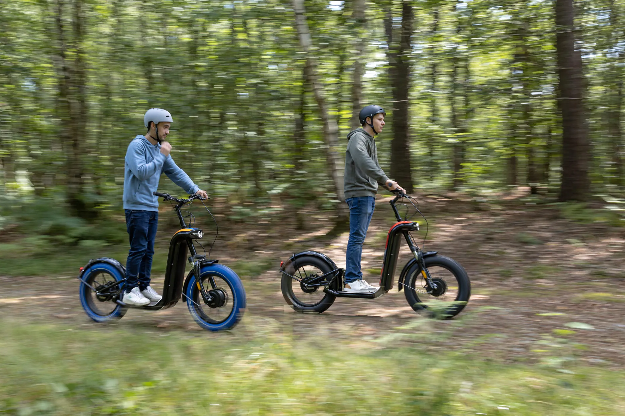 Deux hommes portant des casques roulent sur des trottinettes électriques dans une forêt.