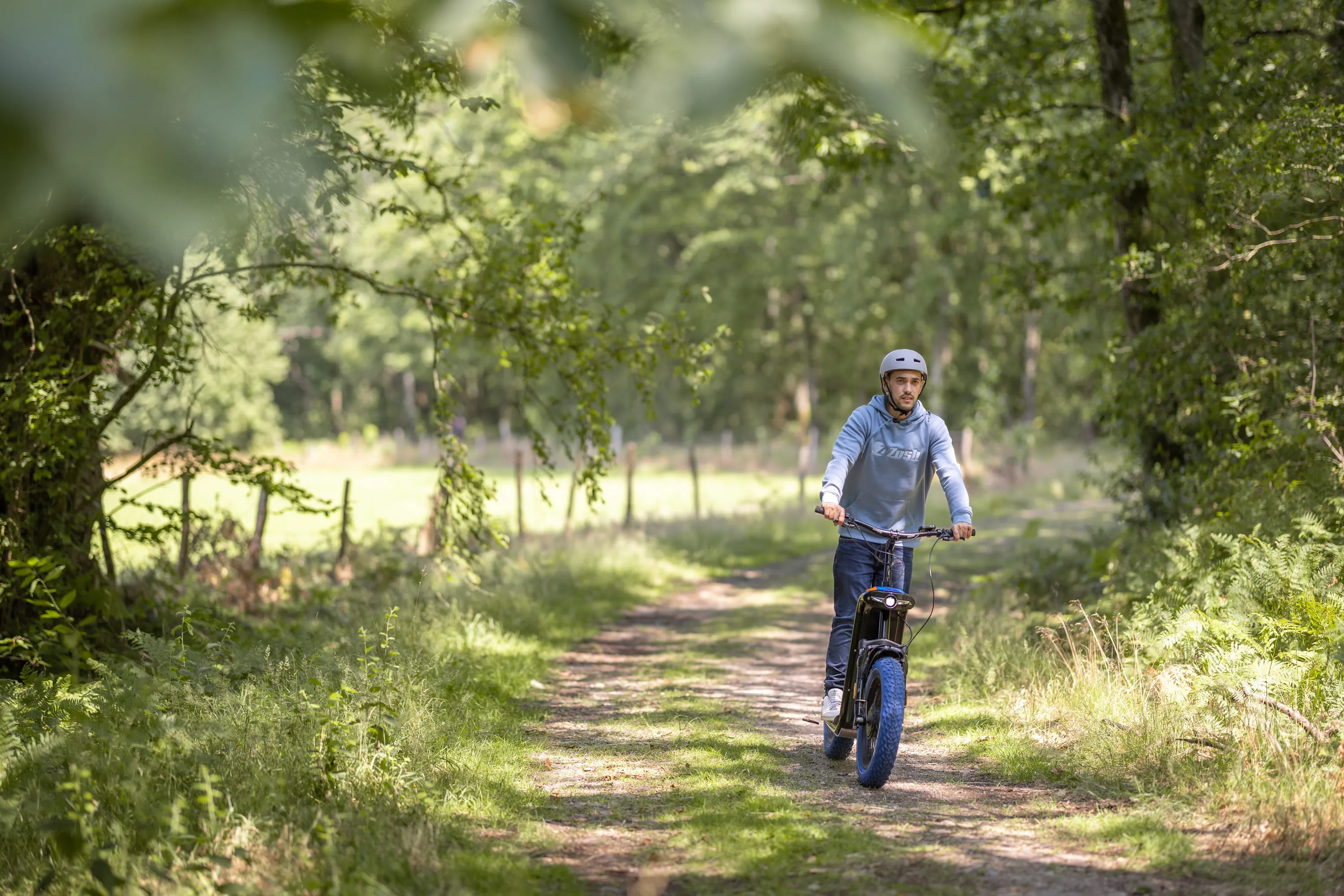 Man wearing helmet riding an electric bike on a dirt path through a green forested area.