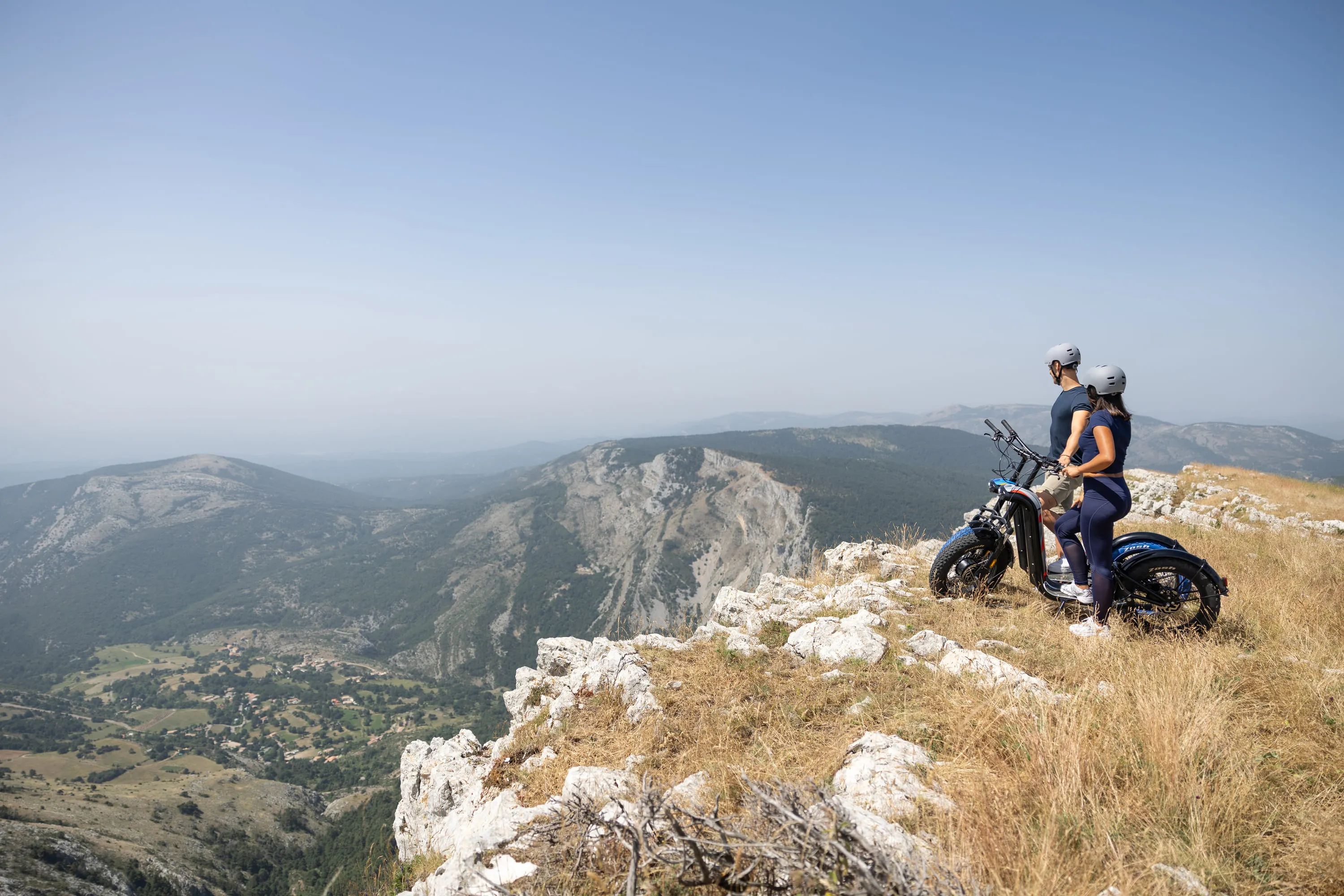 Deux personnes en casques avec vélo électrique regardent une vallée montagneuse depuis un sommet.