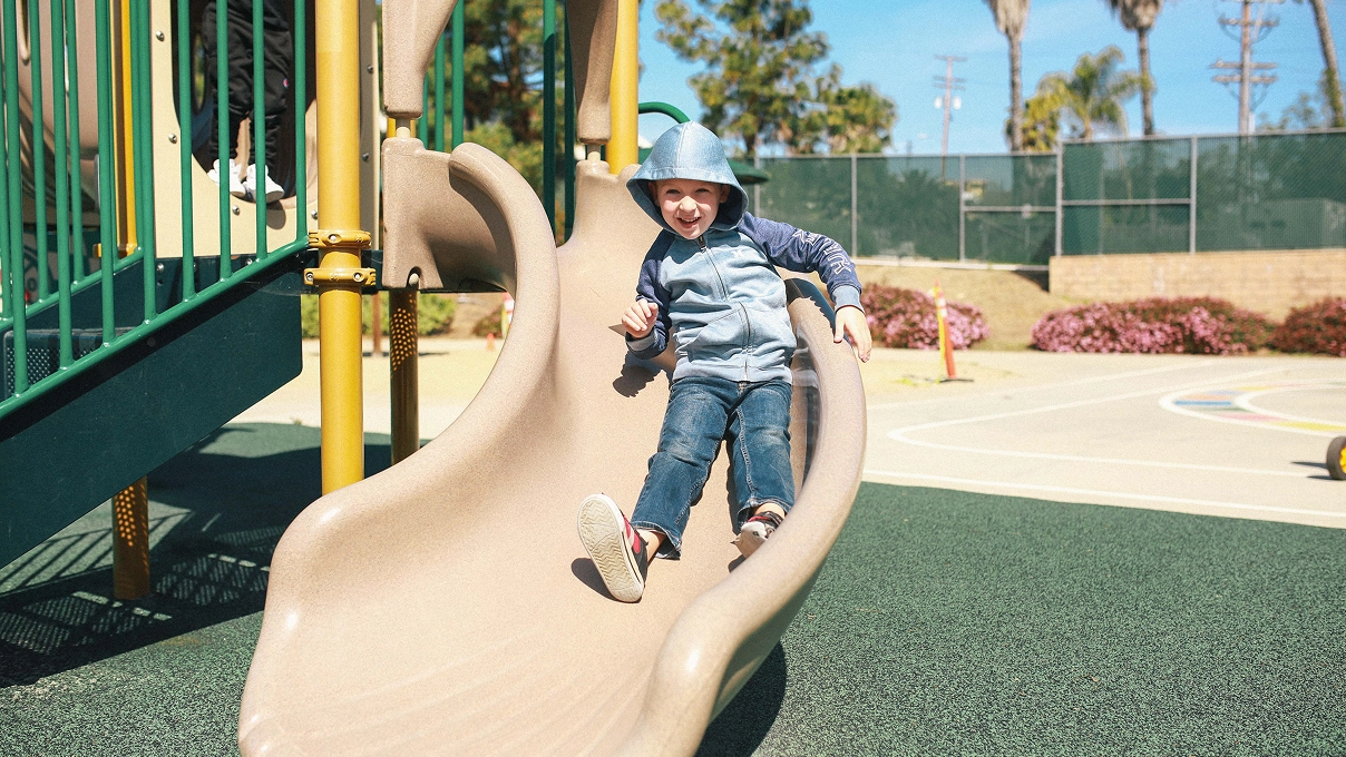 Child wearing a hoodie happily sliding down a tan playground slide on a sunny day.