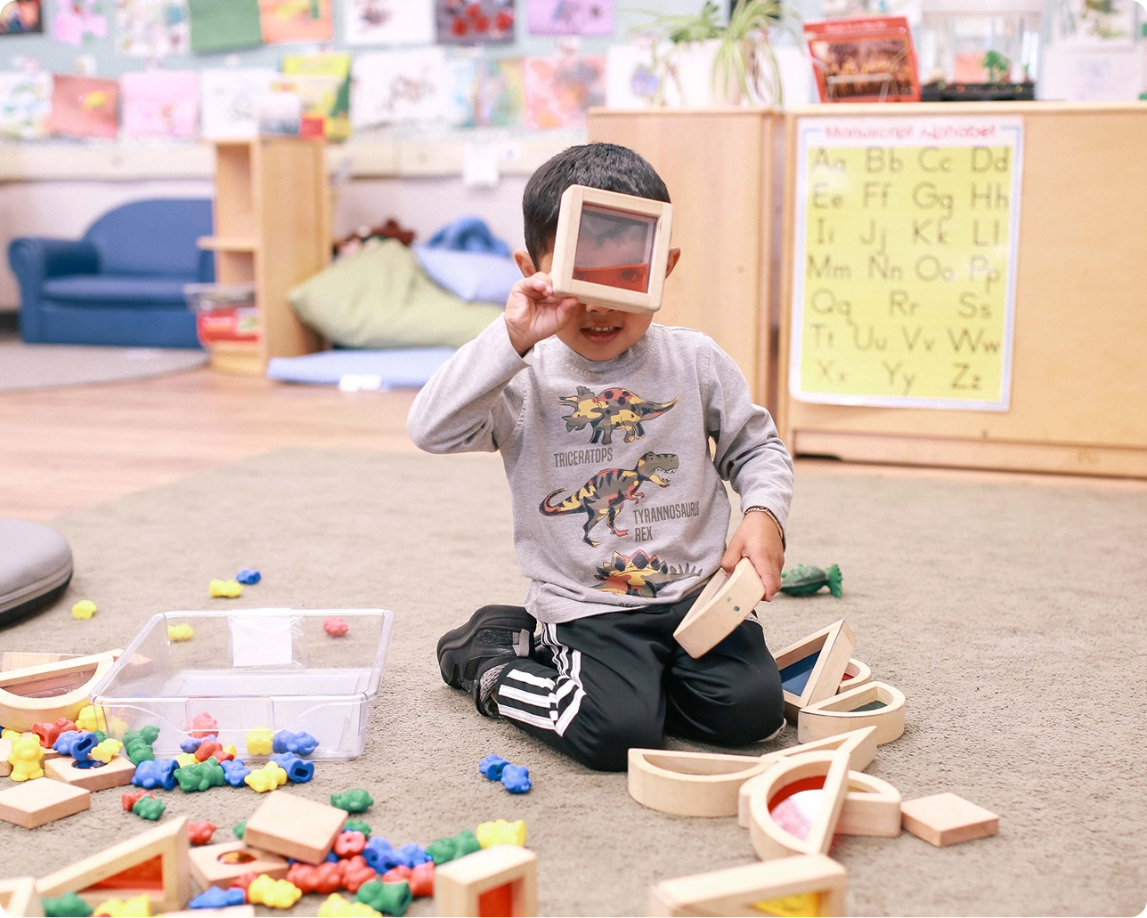 Young boy playing with colored wooden blocks and looking through a translucent red block in a classroom.