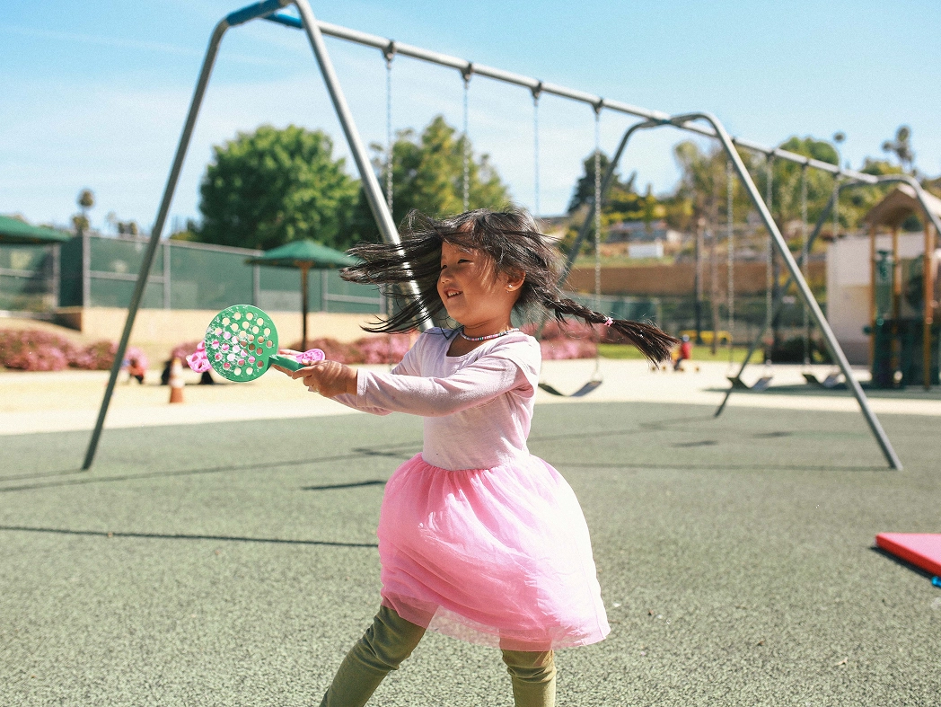 Girl in pink dress playing with a green bubble wand at a sunny playground with swings.