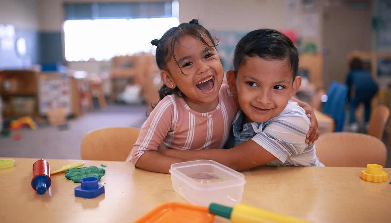 Two young children smiling and hugging while seated at a classroom table with toys and playdough.