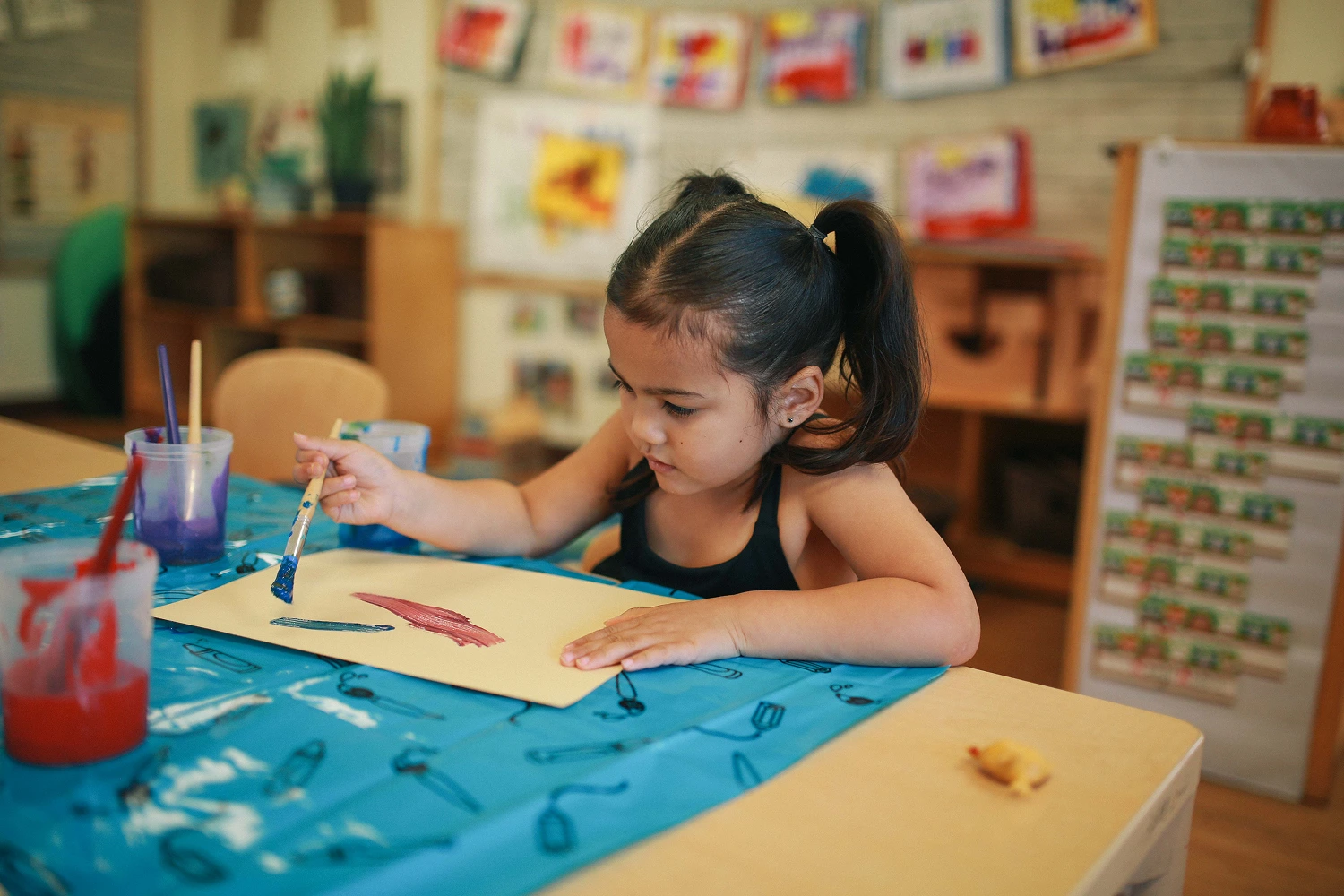 Young girl painting on paper at a table covered with a blue cloth in a classroom.