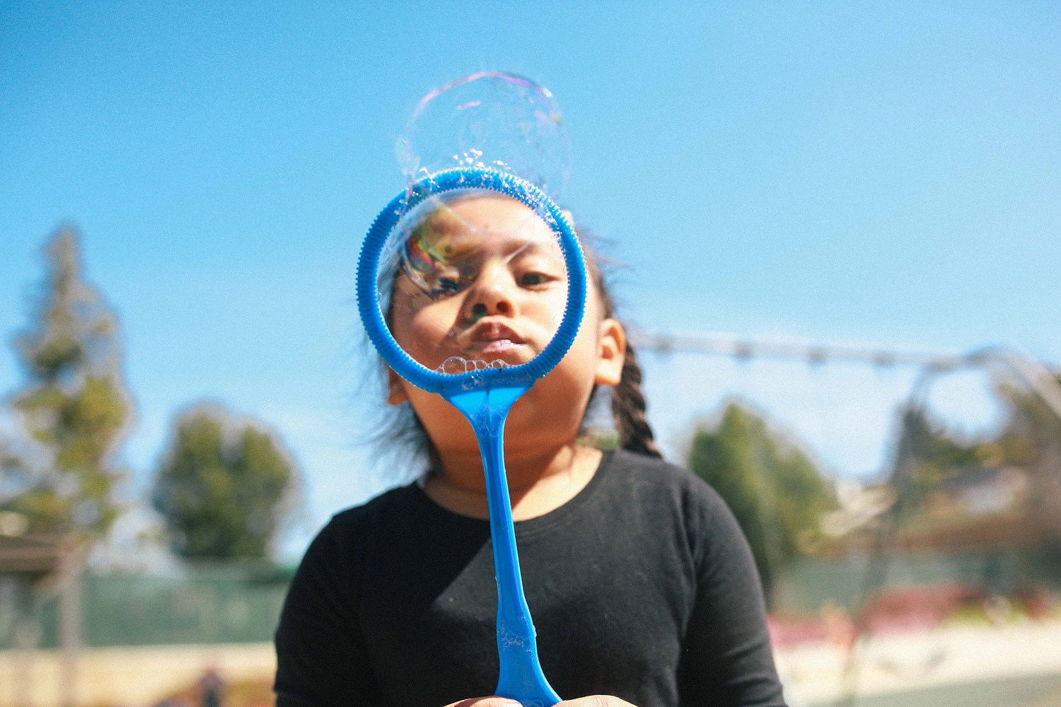 Child blowing soap bubbles outdoors through a large blue bubble wand on a sunny day.