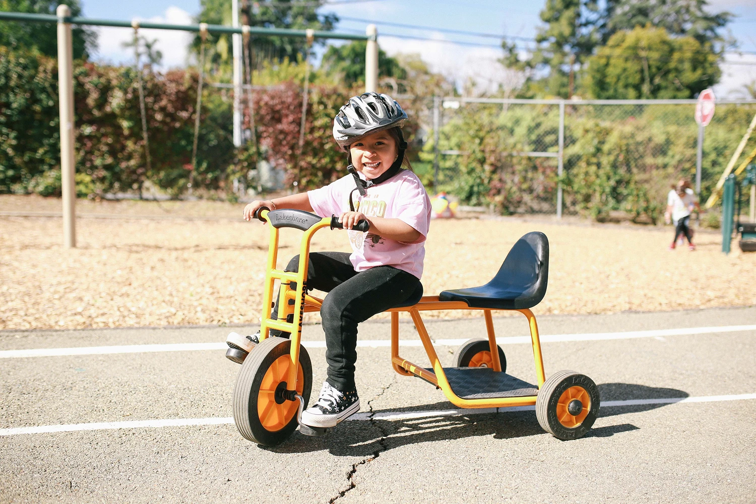Smiling child wearing a helmet rides a large orange tricycle on an outdoor path at a playground.