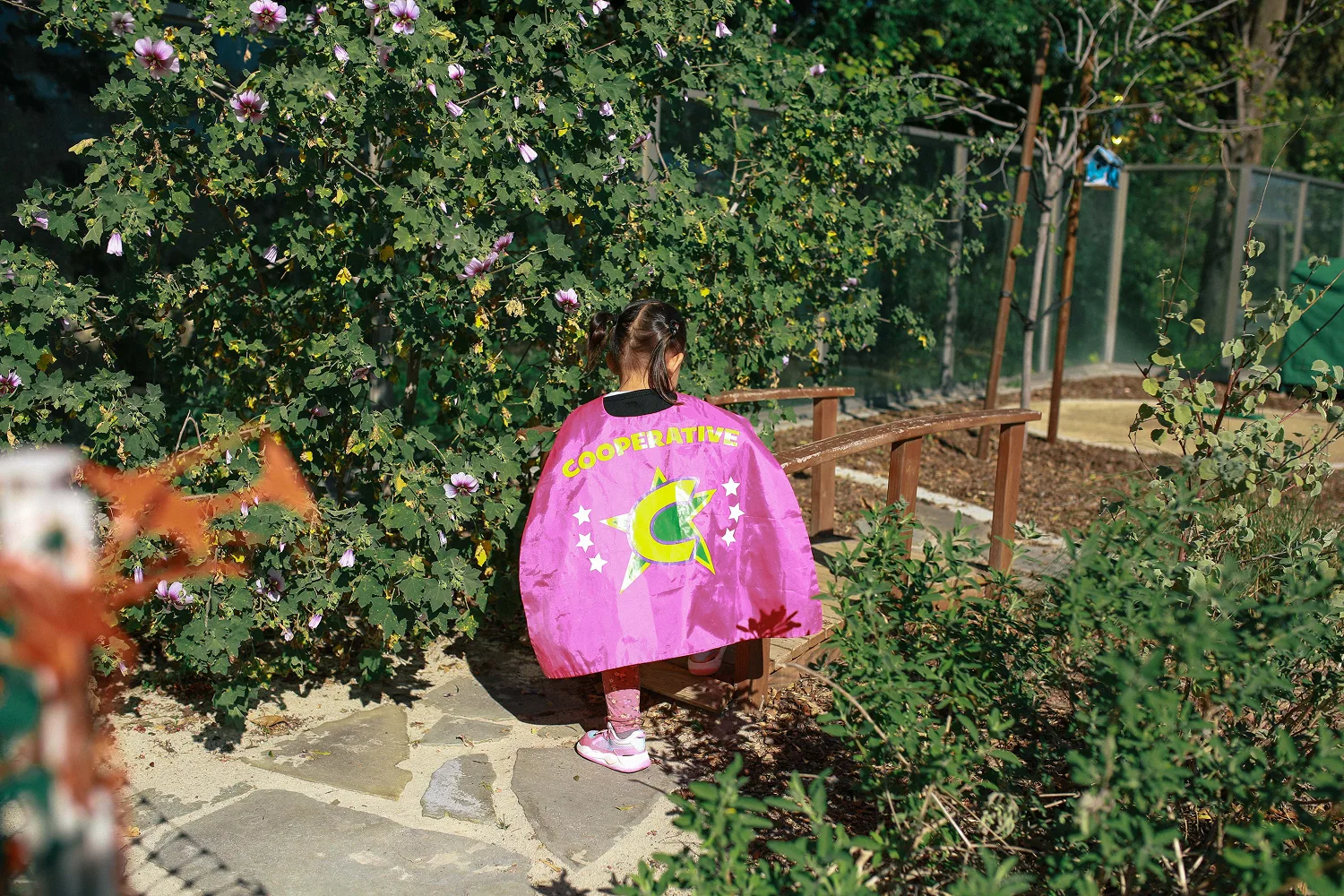 Child with pigtails wearing a pink cape labeled 'Cooperative' walking on a stone path in a garden.
