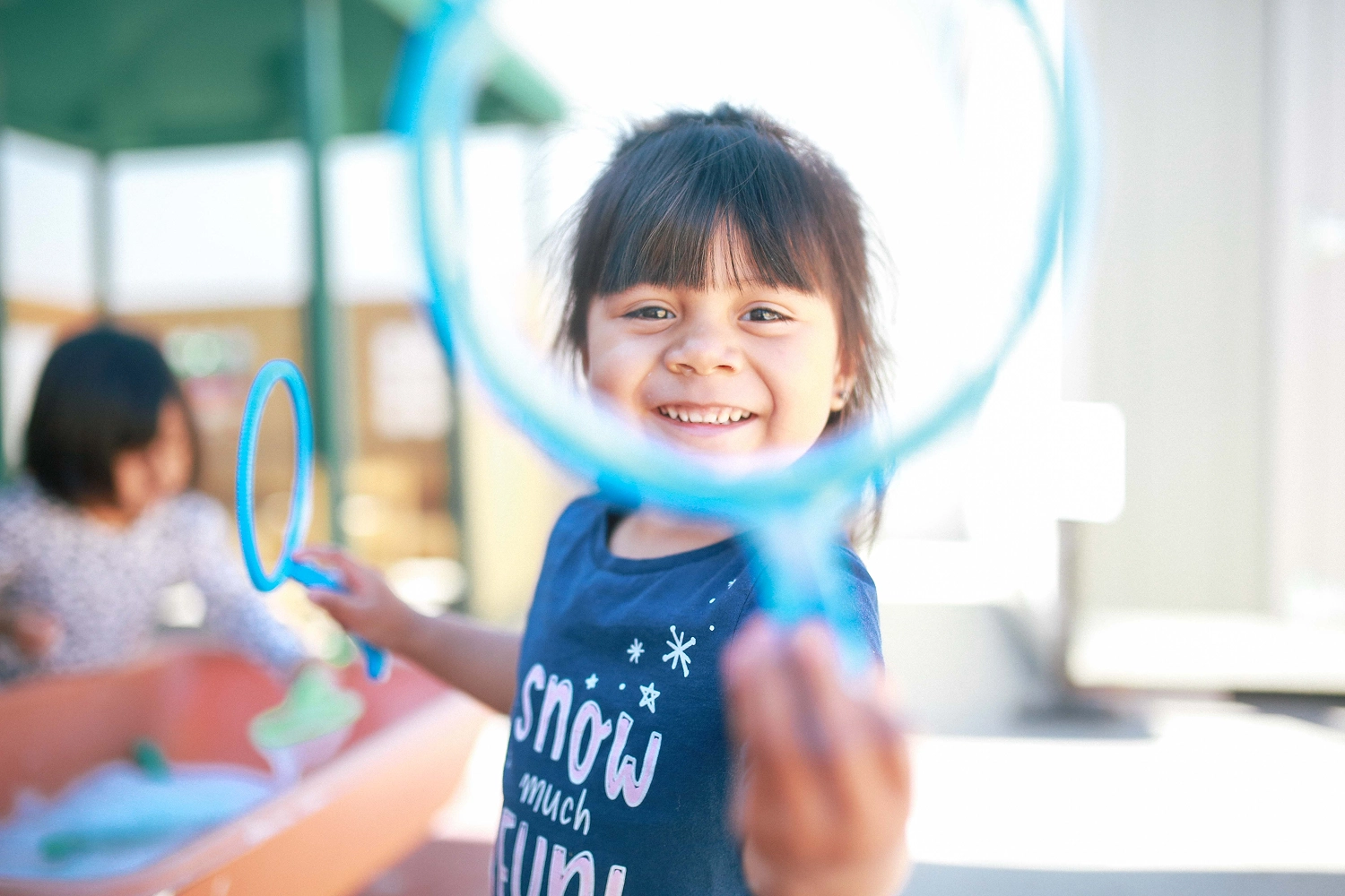 Smiling young girl holding blue bubble wand close to the camera outdoors at a playground.