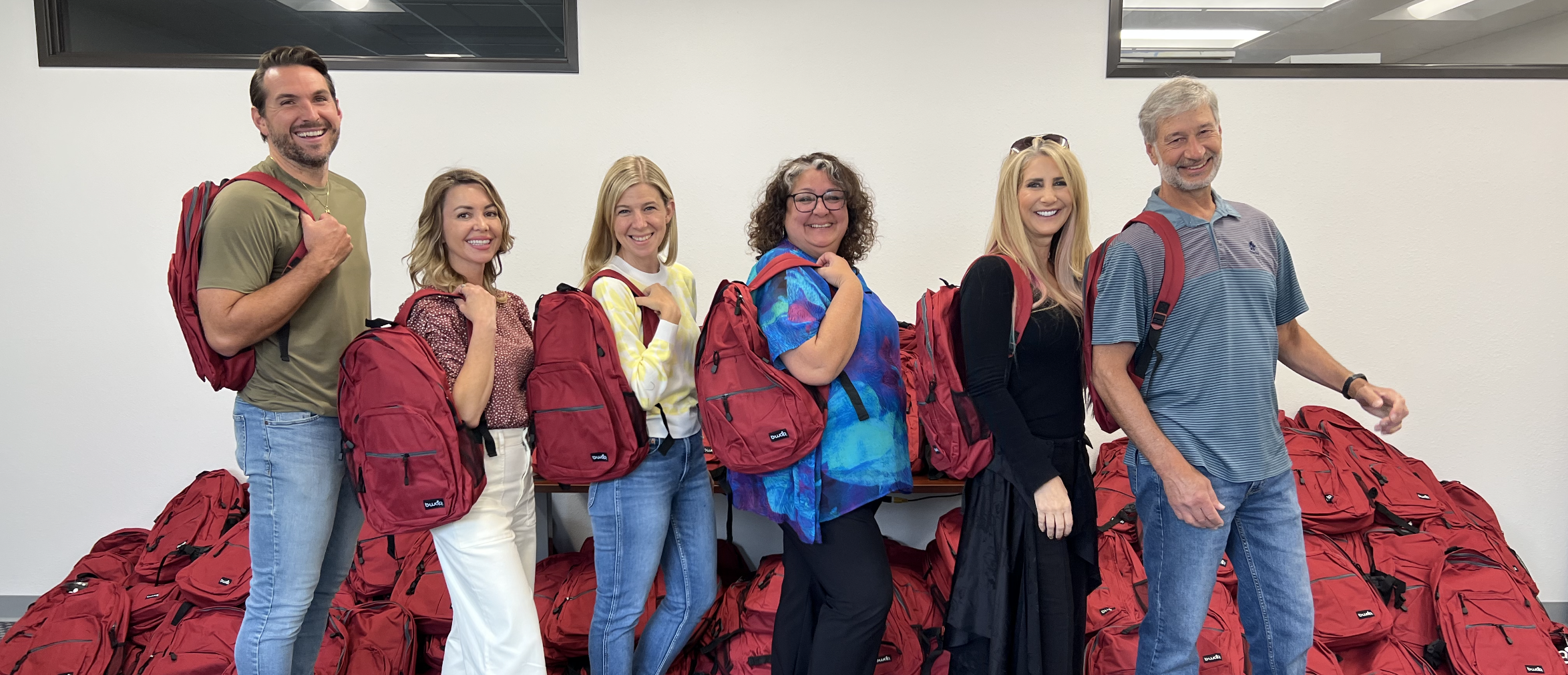 Six smiling adults stand in front of piles of red backpacks, each carrying a red backpack on one shoulder.
