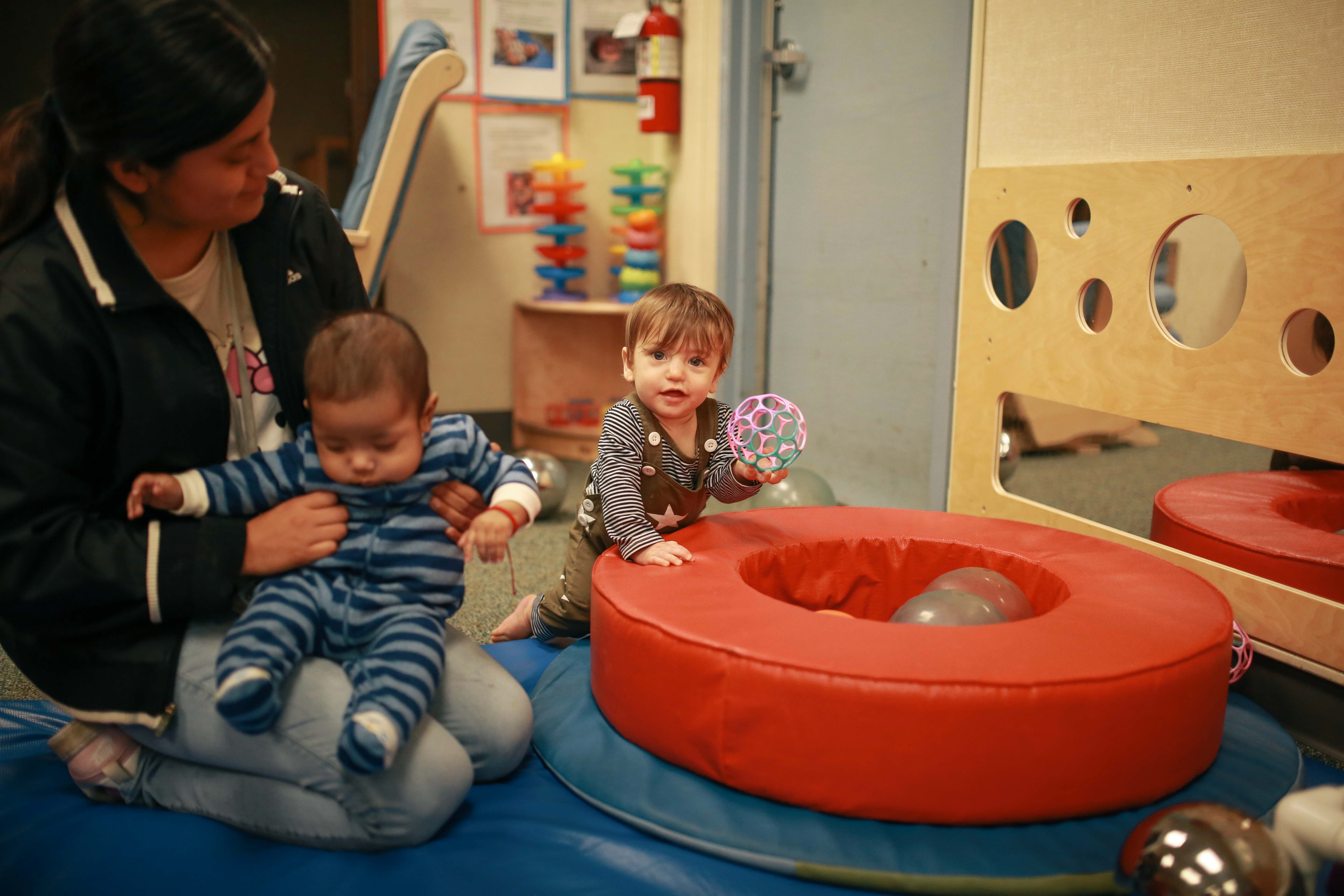 Woman holds baby wearing blue striped outfit while toddler plays with a ball on padded floor.
