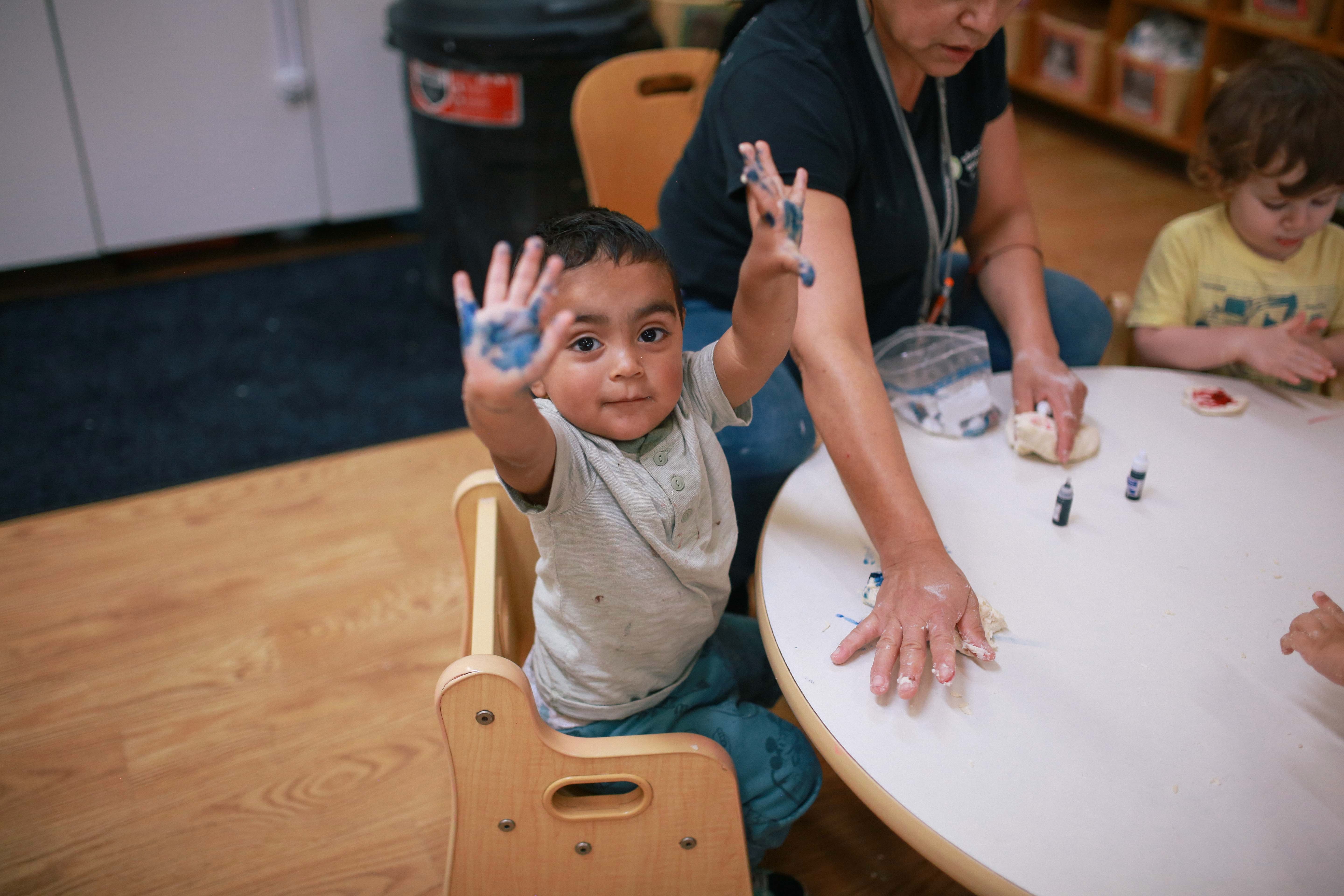 Child with blue paint on hands reaching out, sitting at a table with adults and another child doing crafts.