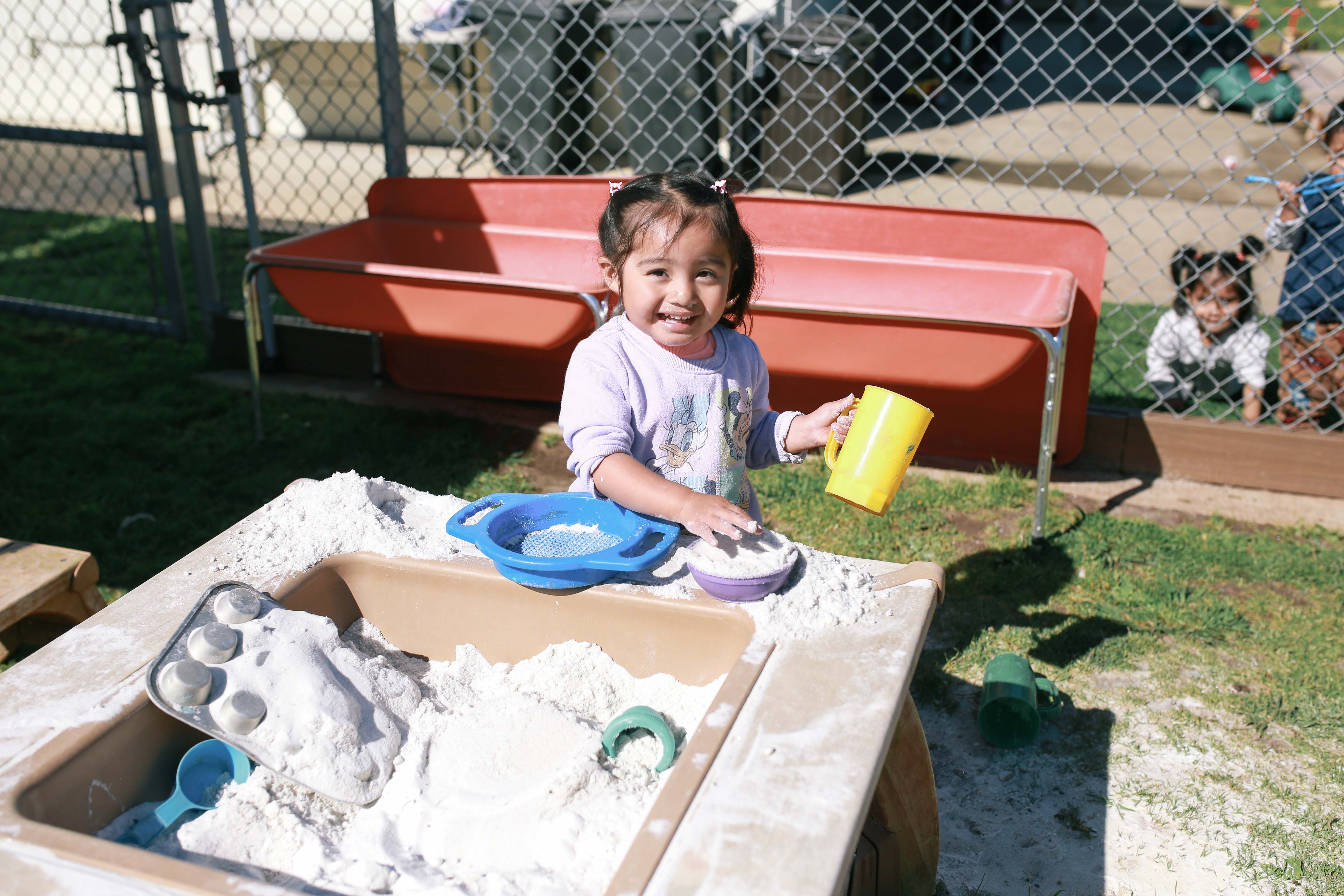Smiling toddler playing with sand in a sandbox holding a yellow cup outdoors.