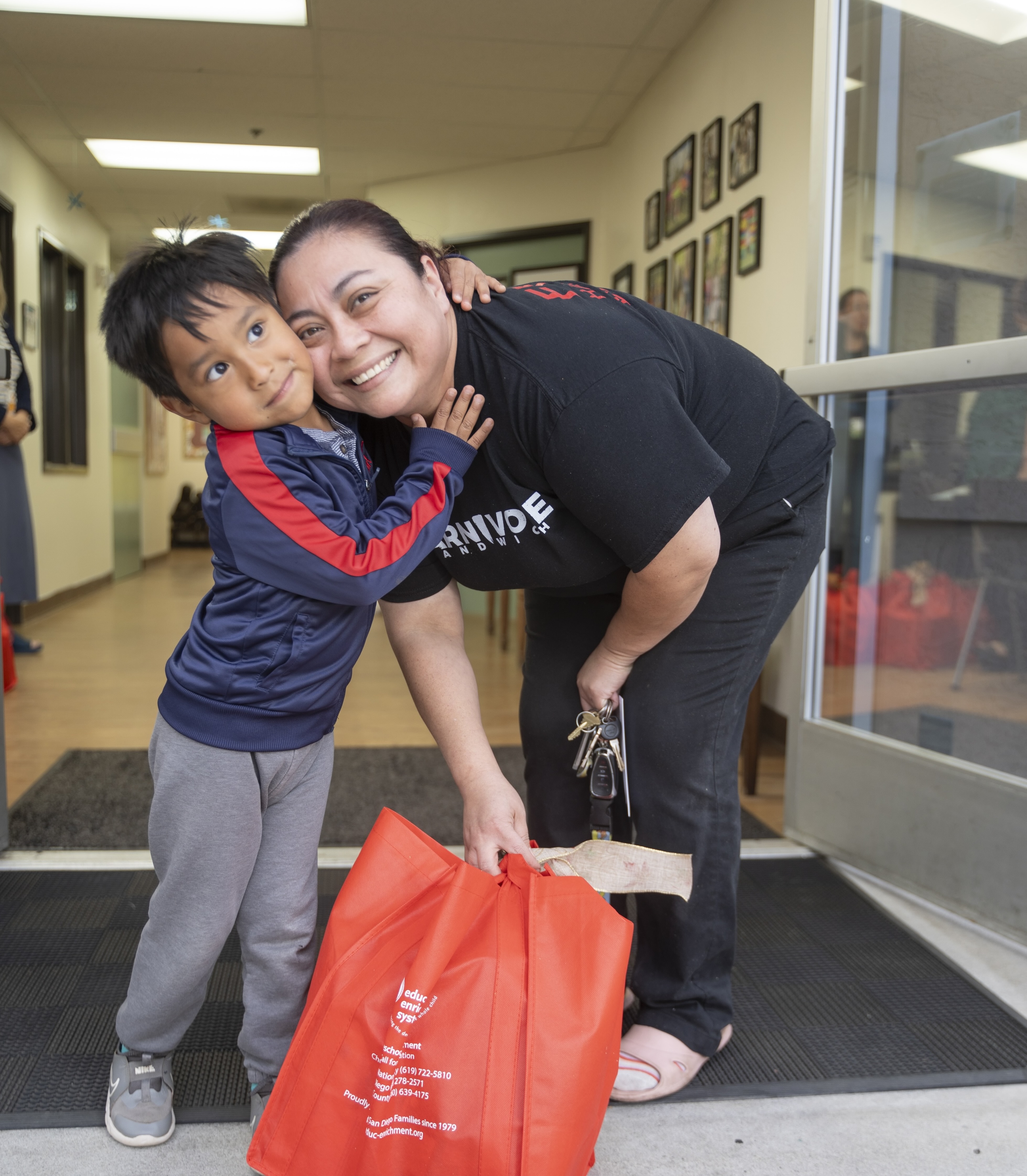 Smiling woman and young boy hugging, woman holding large red reusable bag at building entrance.