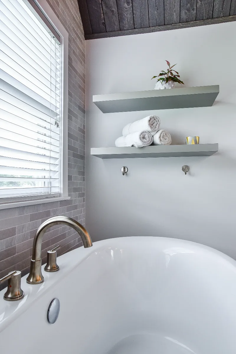 A rustic-warm primary bath in Franklin, TN. Bellmont Alder Folkstone cabinetry with Griege floating shelves, Orion Blanco Mist quartz, Delta Trinsic Champagne Bronze, Kohler Sunstruck tub, and Cassettone Honey Nut wood-look floor tile by Sebring.