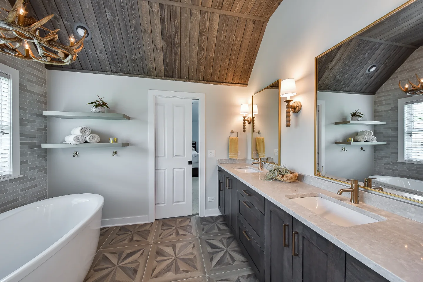 A rustic-warm primary bath in Franklin, TN. Bellmont Alder Folkstone cabinetry with Griege floating shelves, Orion Blanco Mist quartz, Delta Trinsic Champagne Bronze, Kohler Sunstruck tub, and Cassettone Honey Nut wood-look floor tile by Sebring.