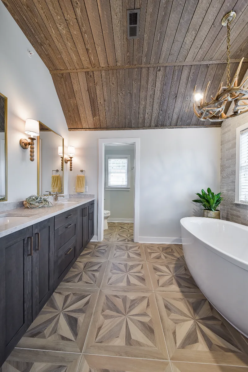 A rustic-warm primary bath in Franklin, TN. Bellmont Alder Folkstone cabinetry with Griege floating shelves, Orion Blanco Mist quartz, Delta Trinsic Champagne Bronze, Kohler Sunstruck tub, and Cassettone Honey Nut wood-look floor tile by Sebring.
