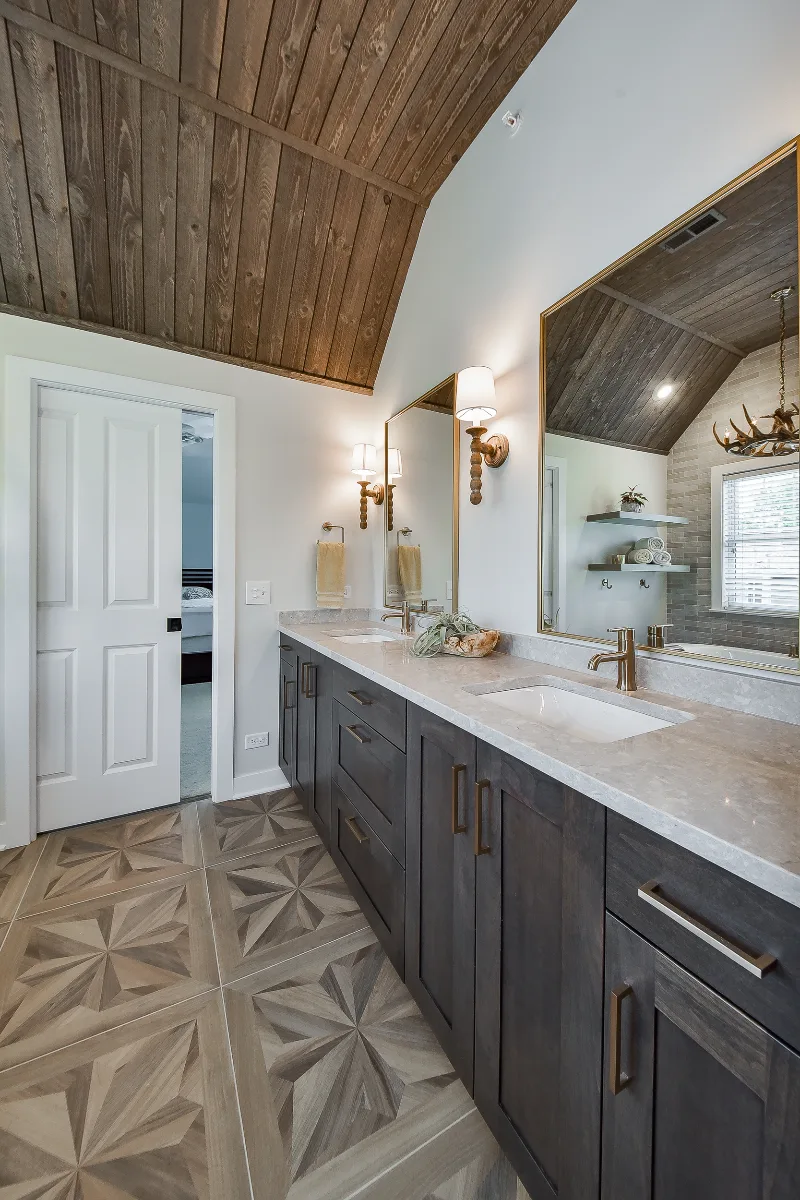 A rustic-warm primary bath in Franklin, TN. Bellmont Alder Folkstone cabinetry with Griege floating shelves, Orion Blanco Mist quartz, Delta Trinsic Champagne Bronze, Kohler Sunstruck tub, and Cassettone Honey Nut wood-look floor tile by Sebring.