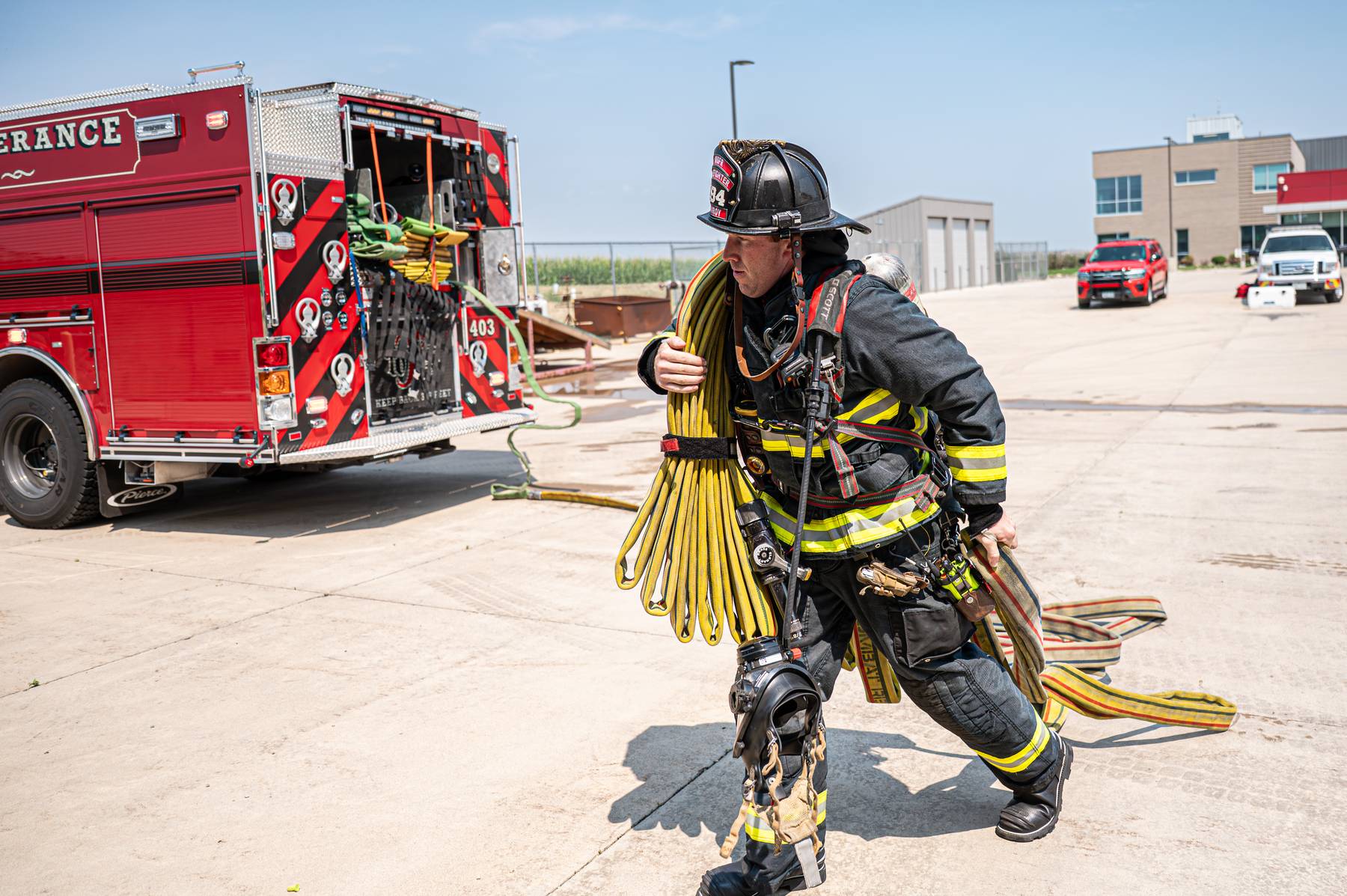 Firefighter in full gear carrying coiled hoses running near a red fire truck on a concrete surface.