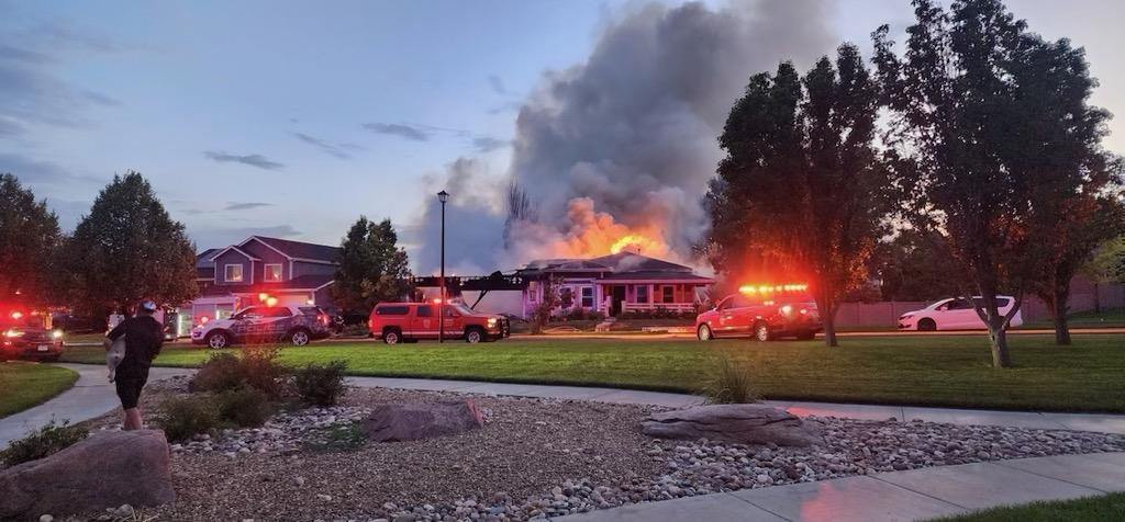 House engulfed in flames with fire trucks and smoke rising at dusk in a suburban neighborhood.
