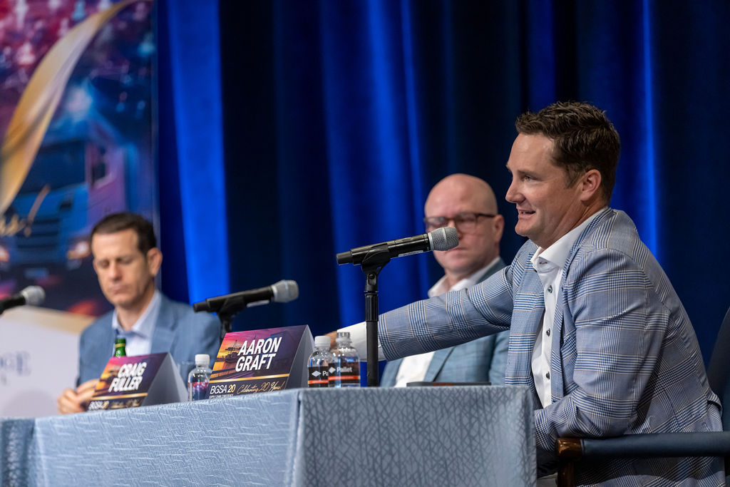 Three men in suits at a panel discussion with microphones and nameplates, one man speaking.