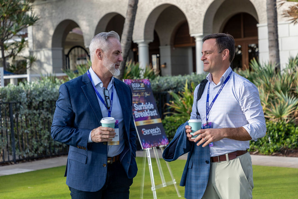 Two men in business attire holding coffee cups and talking outdoors near a sign reading Surfside Breakfast.