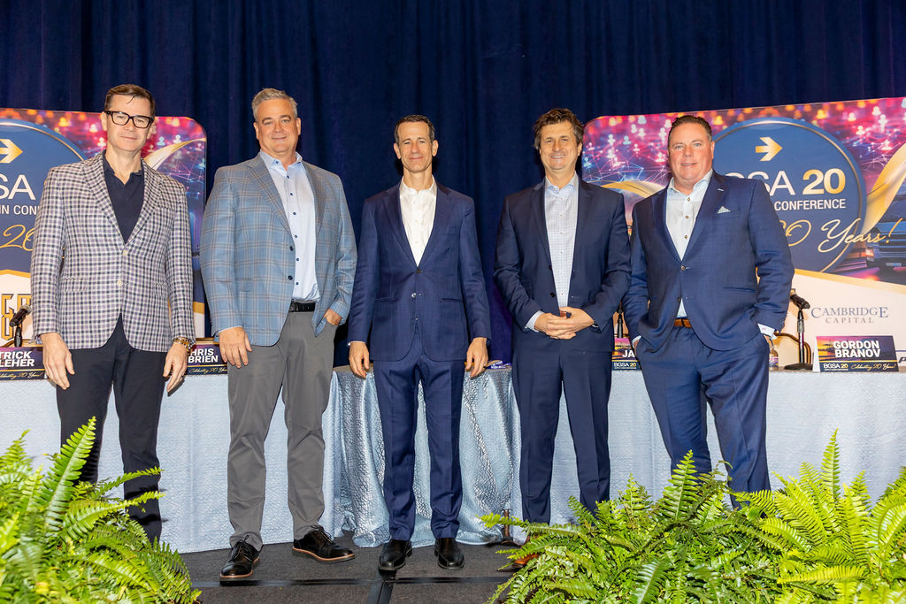 Five men in business attire standing in front of a conference stage with banners and plants.
