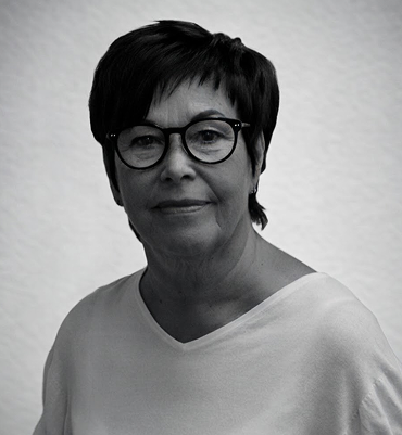 Leende äldre kvinna med kort mörkt hår och glasögon mot ljus bakgrund. LEKRI Sandviken Lena