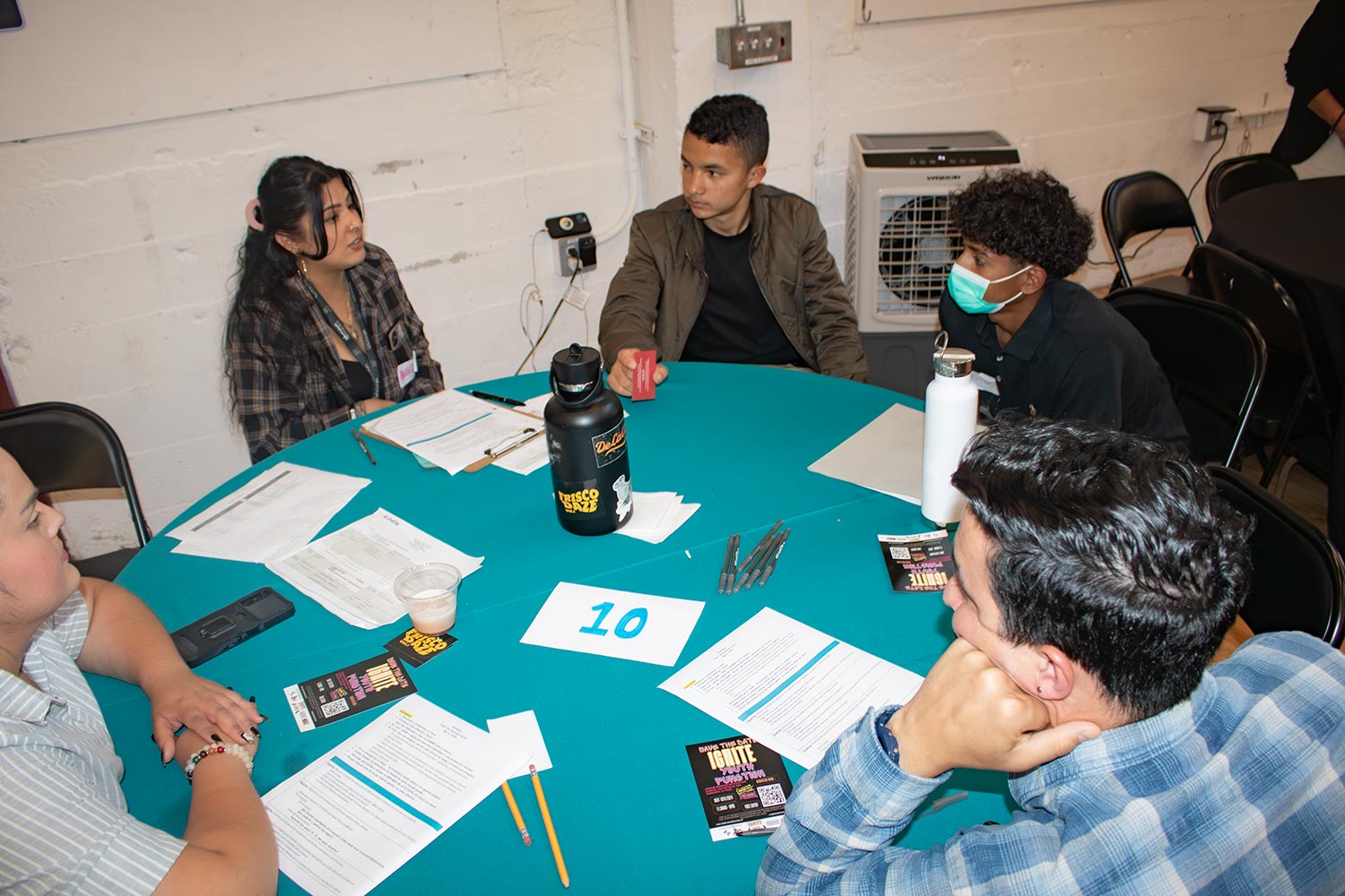 Five people seated around a table engaged in discussion with papers, pens, and water bottles on the table.