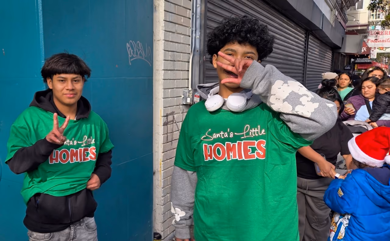 Two young males wearing green Santa's Little Homies shirts posing with peace signs on a city sidewalk.