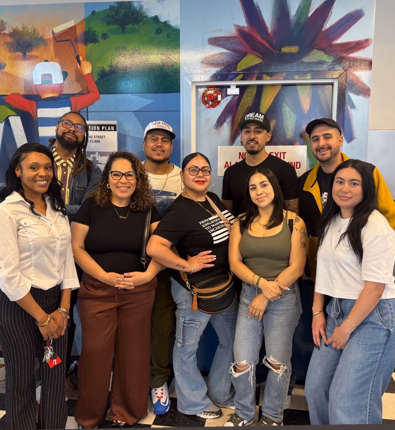 Group of nine diverse people smiling and posing indoors against a colorful mural background.
