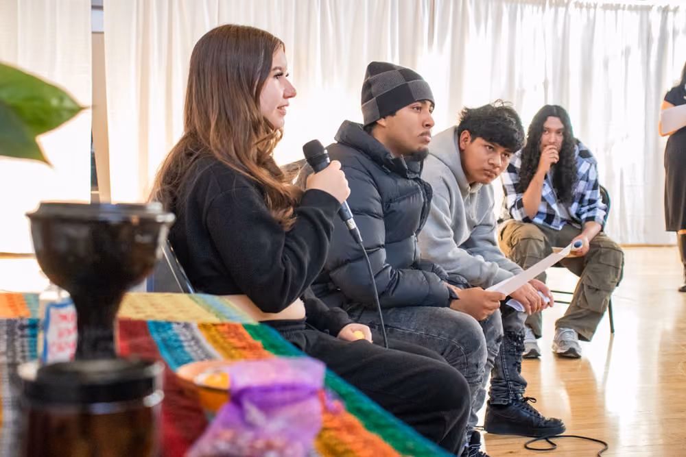 Four young people seated indoors; one woman speaks into a microphone; others listen attentively.