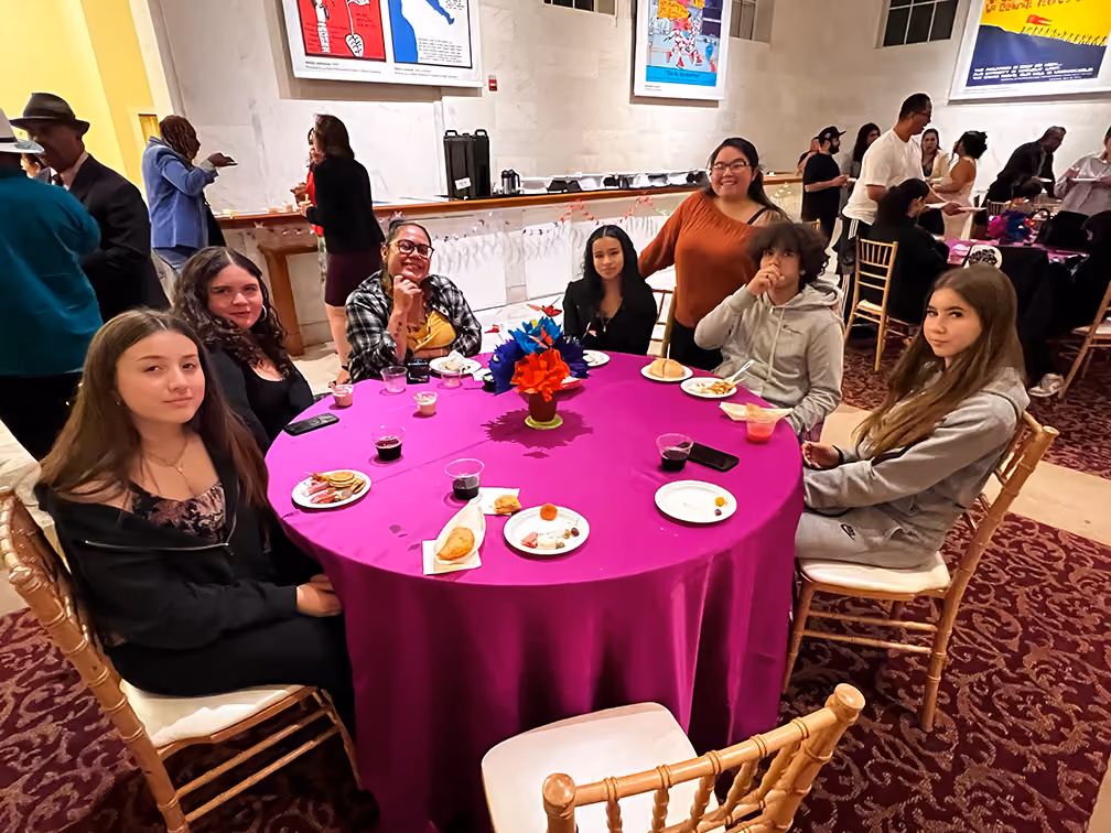 Seven young people seated around a purple tablecloth with snacks and drinks at an indoor event.