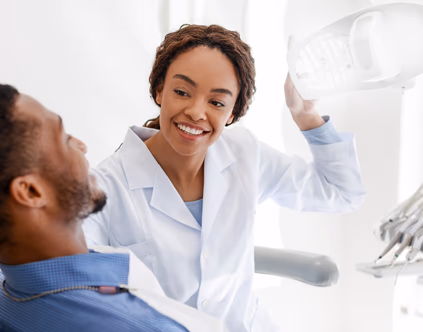 Smiling female dentist in white coat adjusting a dental X-ray machine while patient sits in chair.