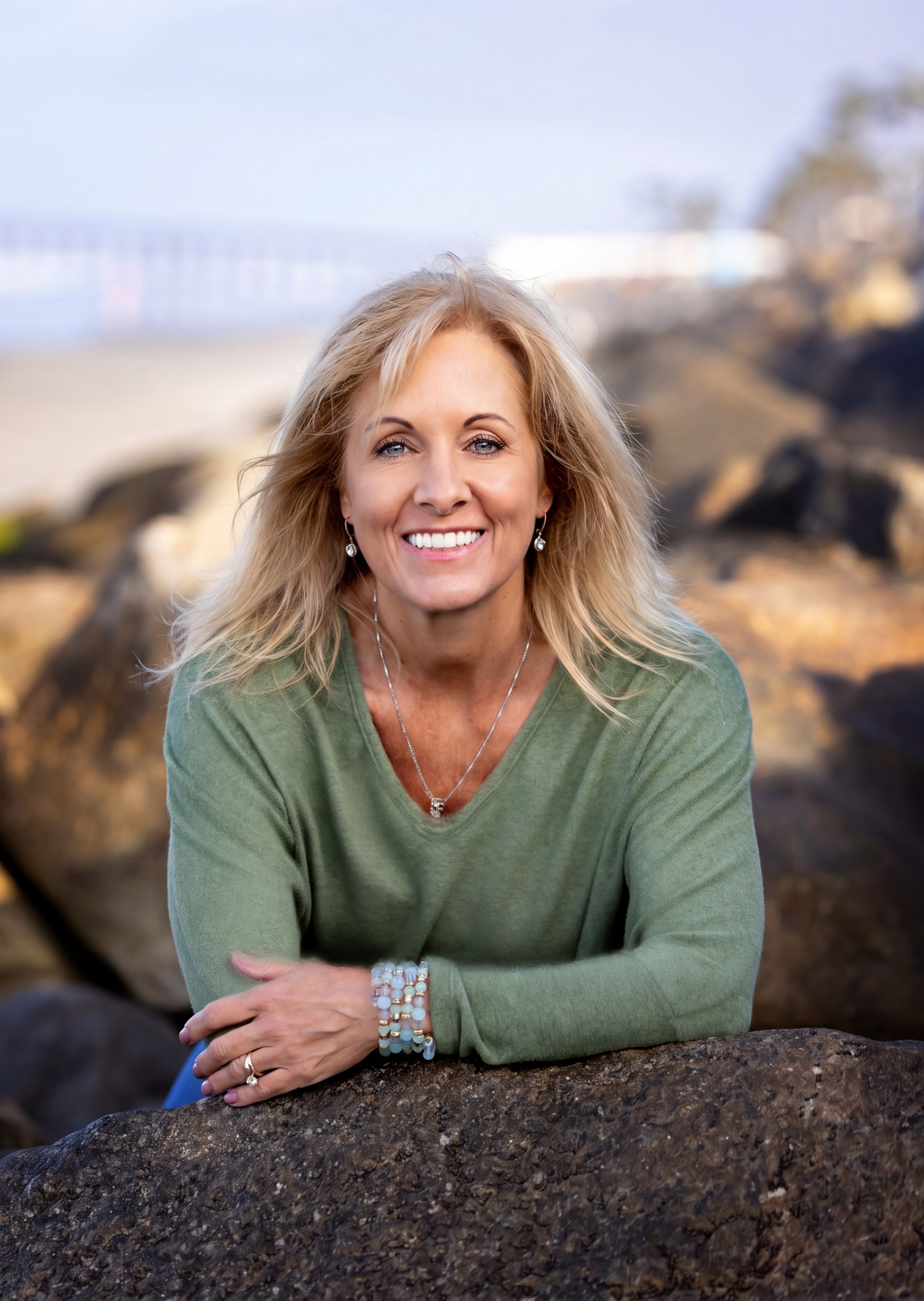 Susie smiling in a green sweater leaning on a large rock outdoors with blurred background.
