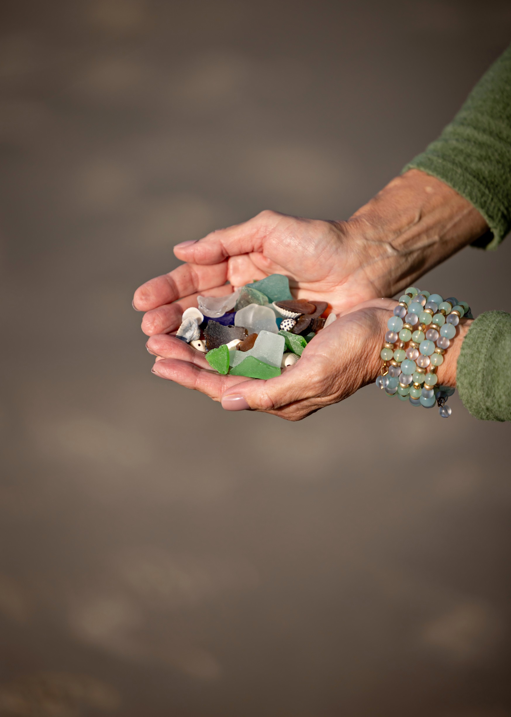 Hands holding assorted sea glass pieces and small shells against a blurred background.