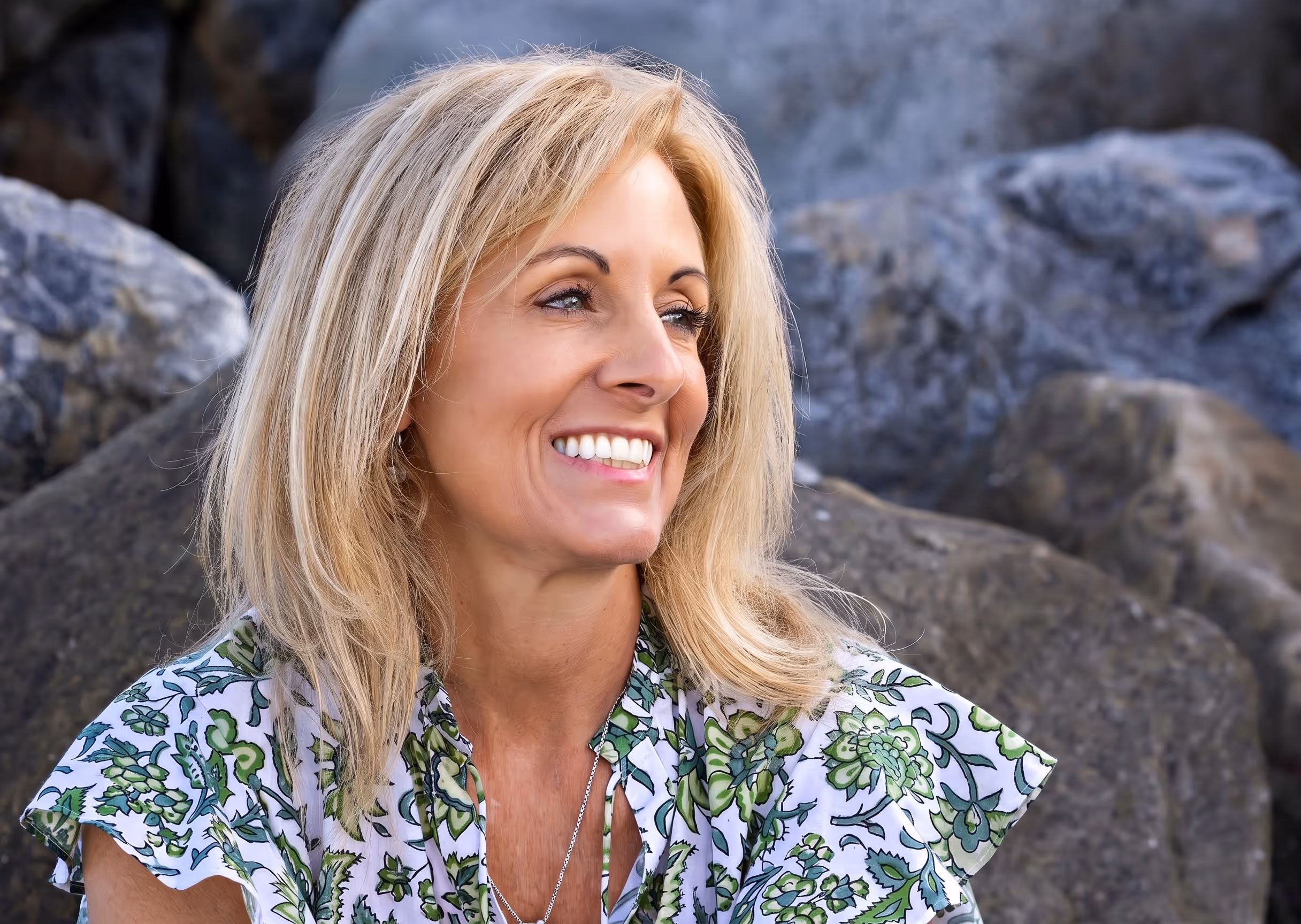 Susie Valencia wearing a green floral top sitting outdoors with large gray rocks behind her.