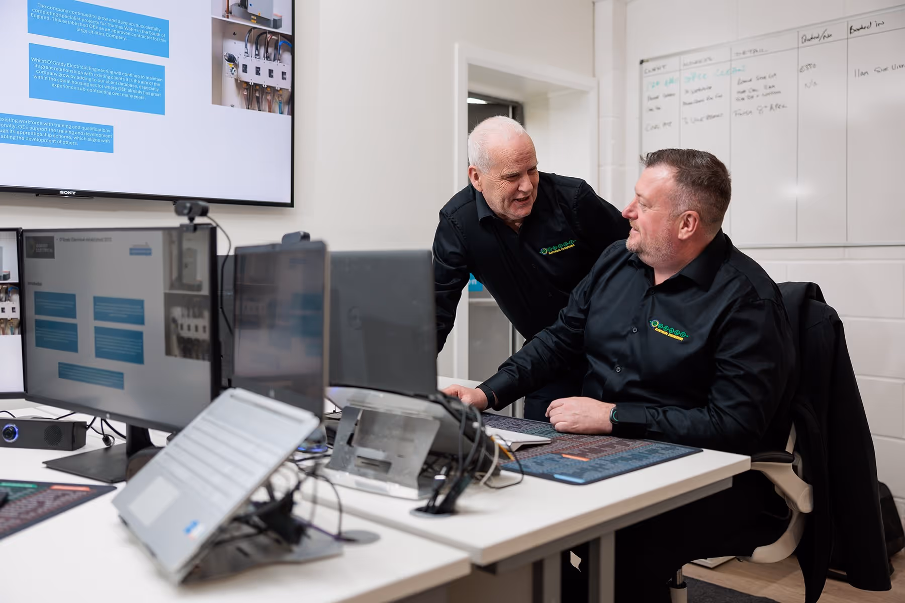 O'Grady management team working in the office. One man is sat at his desk talking to another man who is stood beside him in the office.