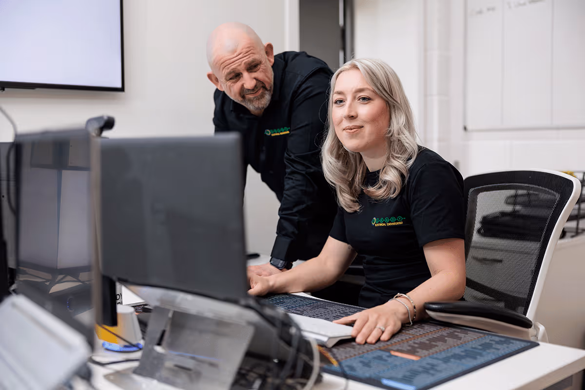 O'Grady management team working in the office. A woman is sitting down at her desk looking at the screen. A man is looking over her shoulder reviewing the screen with her.