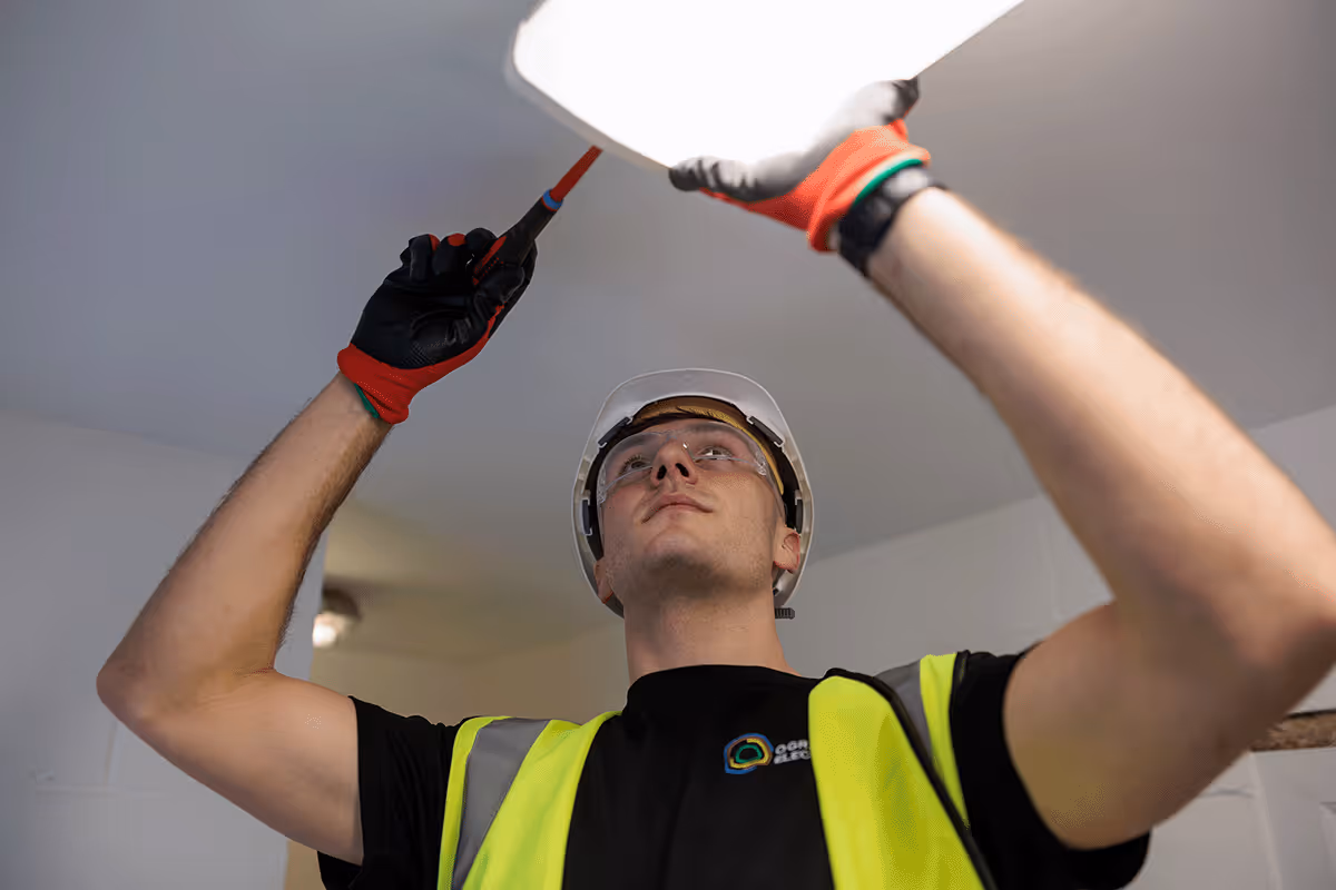 An O'Grady team member in uniform standing on a ladder working on a building lighting system. He is testing the light fitting.