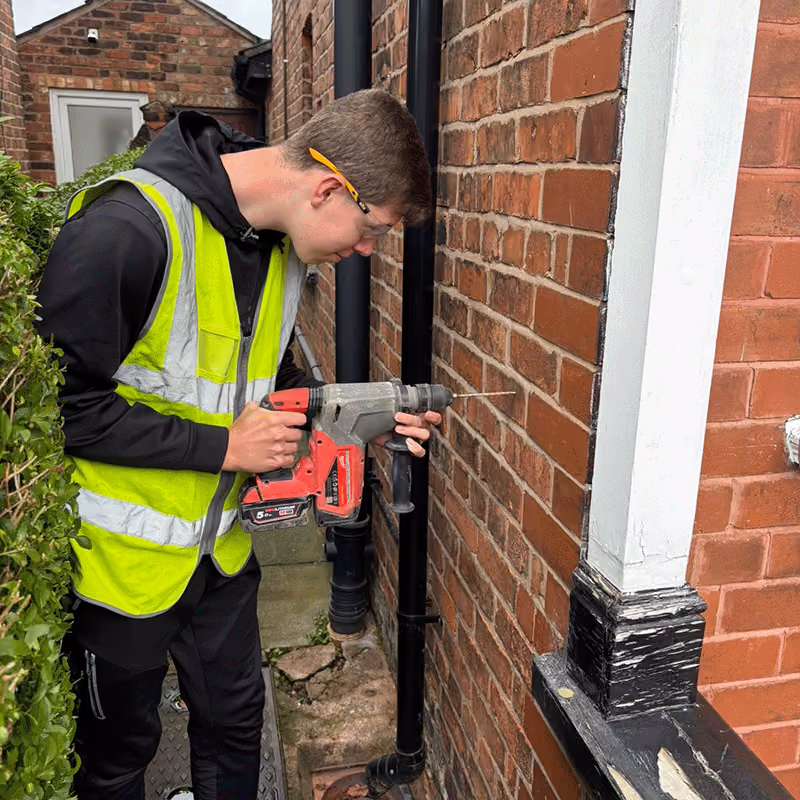 A team member wearing a yellow safety vest uses a red and gray power drill to drill into a brick wall outside a red brick building.