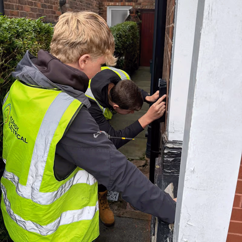 Two team members wearing high-visibility vests working on an electrical installation outside a brick building, with one holding a tool and the other adjusting a black fixture on the wall.